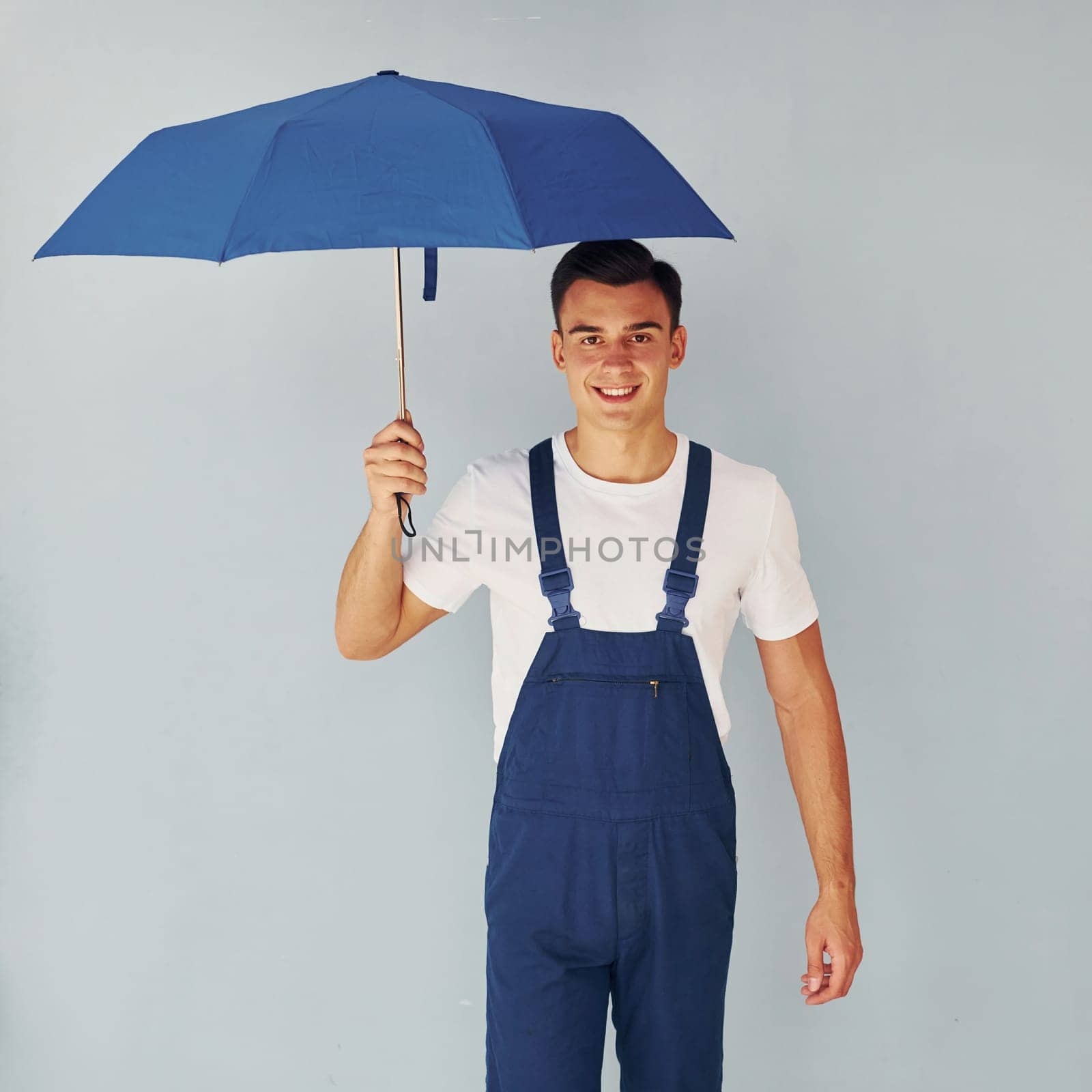 Holds umbrella by hand. Male worker in blue uniform standing inside of studio against white background.