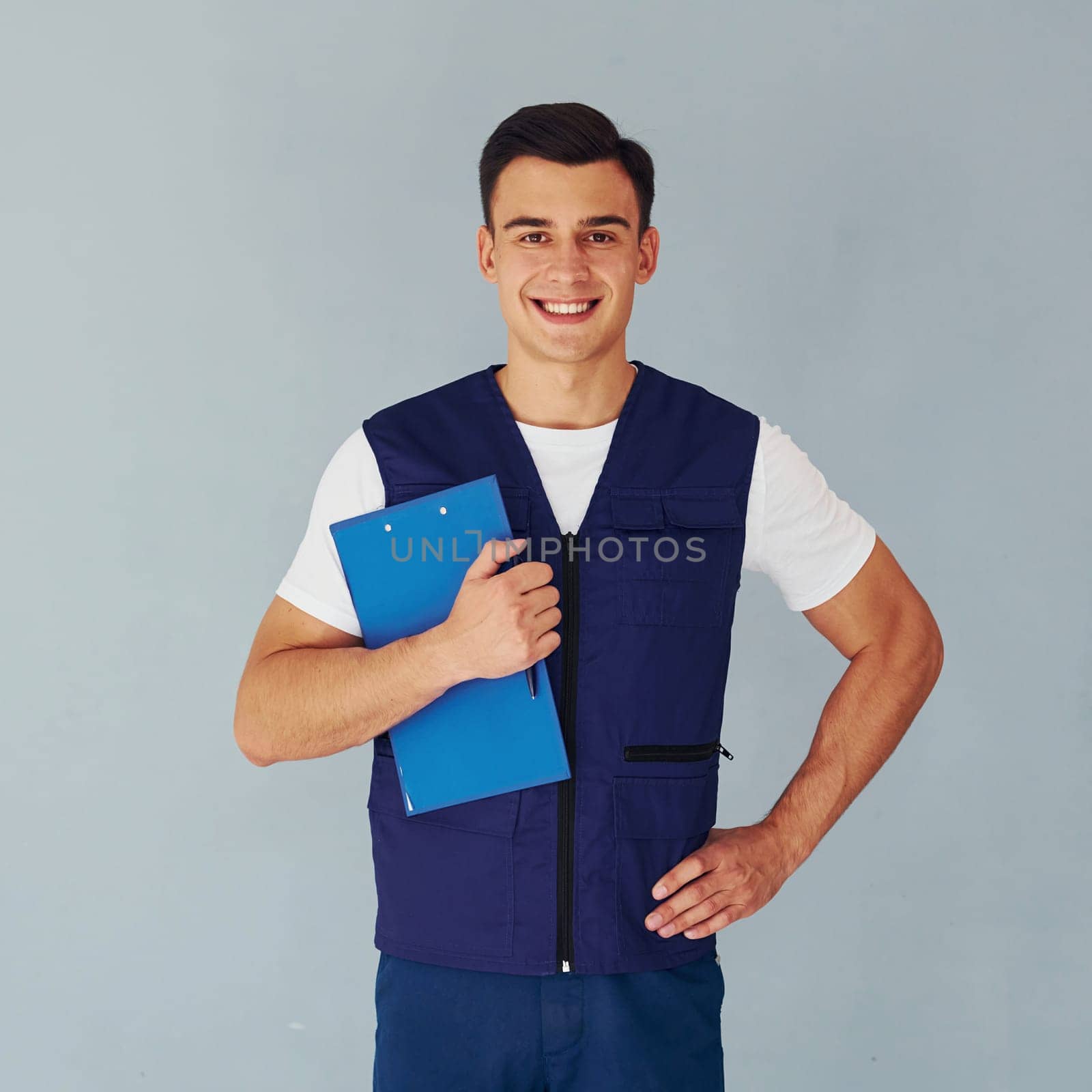 Holds notepad. Male worker in blue uniform standing inside of studio against white background.