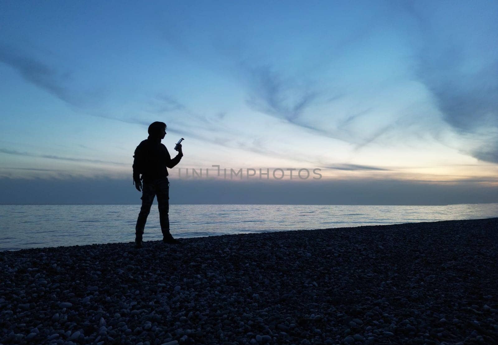 the silhouette of a guy at a beautiful sunset against the background of the sea and clouds in his hand is a remote control from a drone by Rotozey