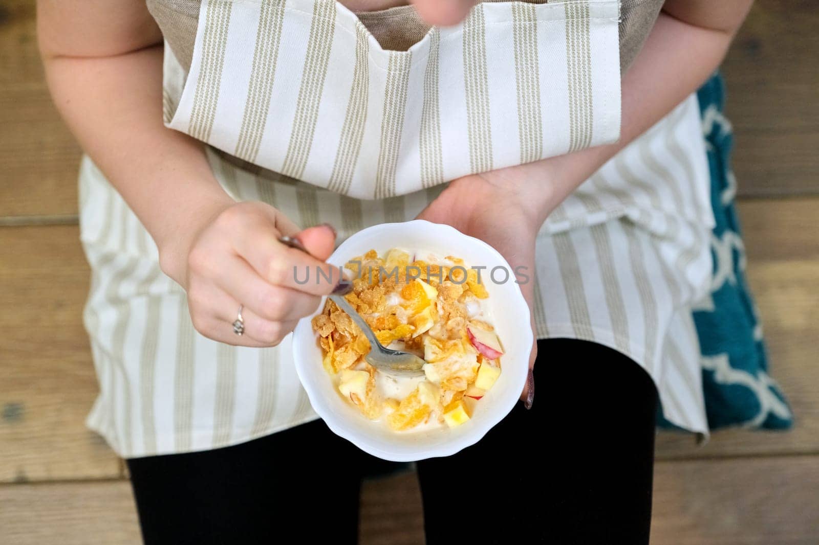Close-up of plate with fruit salad in hands of eating woman sitting at home in kitchen on floor in apron. Healthy diet, vitamin homemade delicious food, corn flavored yogurt fruit salad