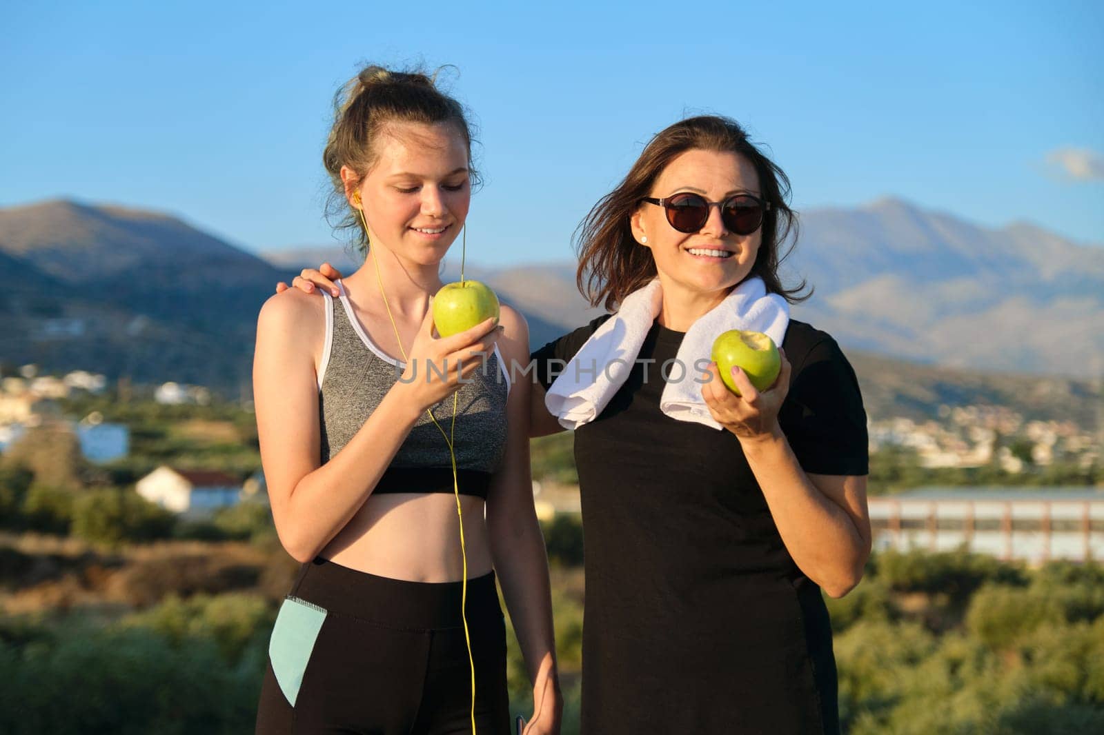Healthy lifestyle and healthy food concept. Smiling fitness mother and teen daughter together eating apples, talking, resting after athletic running and outdoor exercise