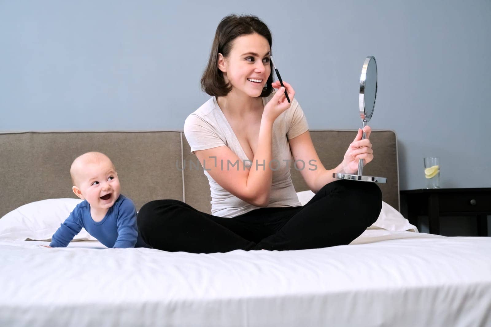 Young beautiful happy mother with toddler son at home in bed, woman with cosmetic mirror does makeup