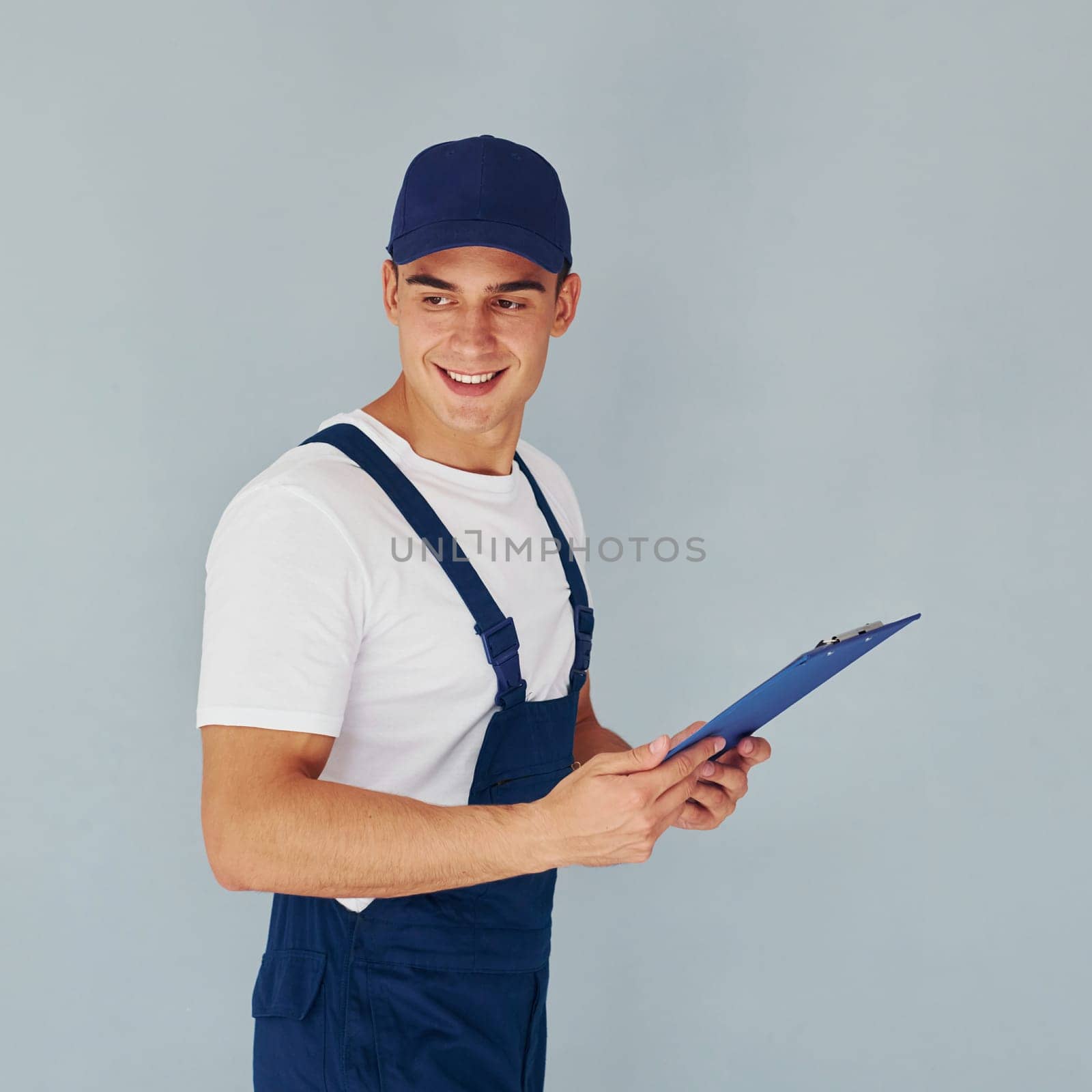 In cap and with notepad. Male worker in blue uniform standing inside of studio against white background.