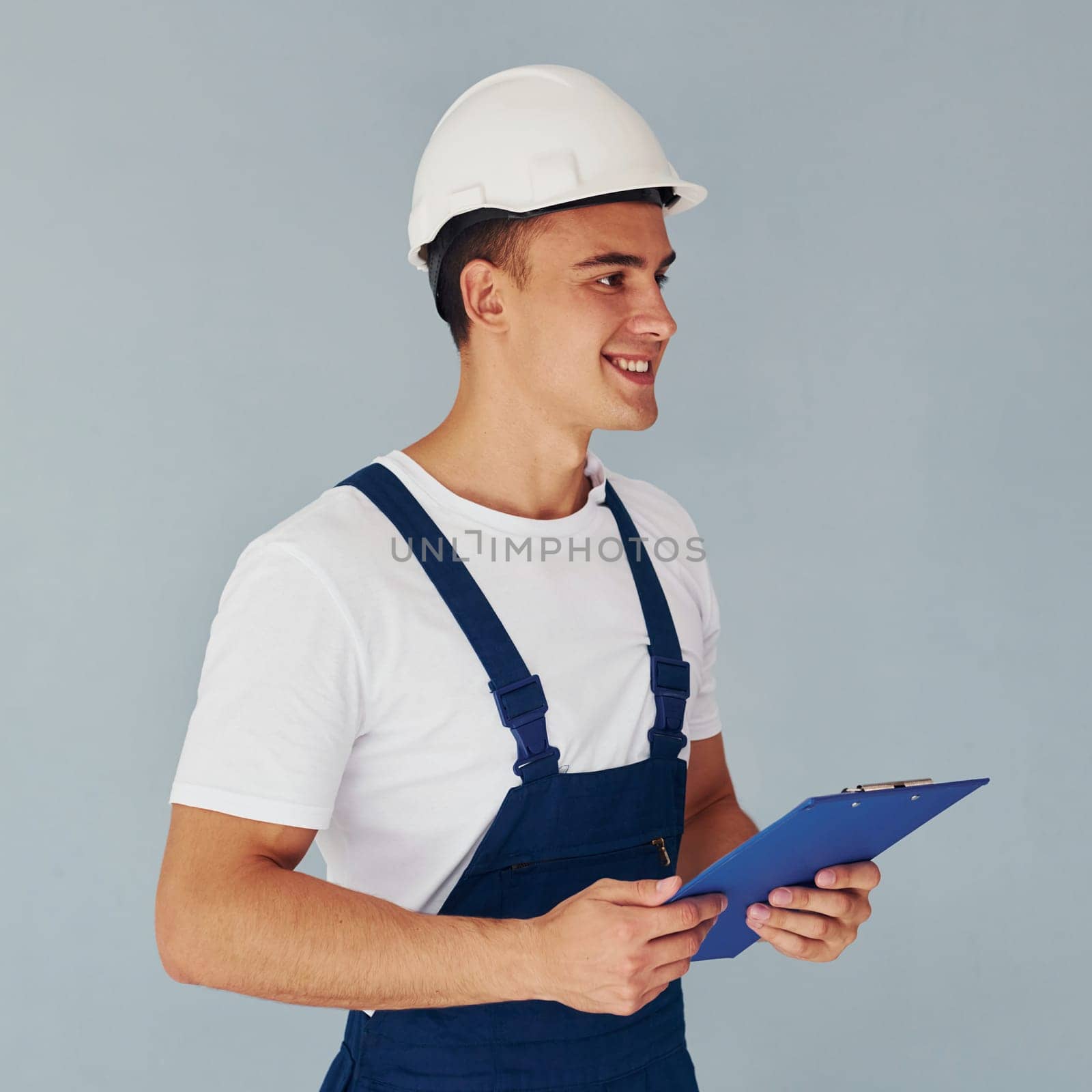 With notepad and hard hat. Male worker in blue uniform standing inside of studio against white background.