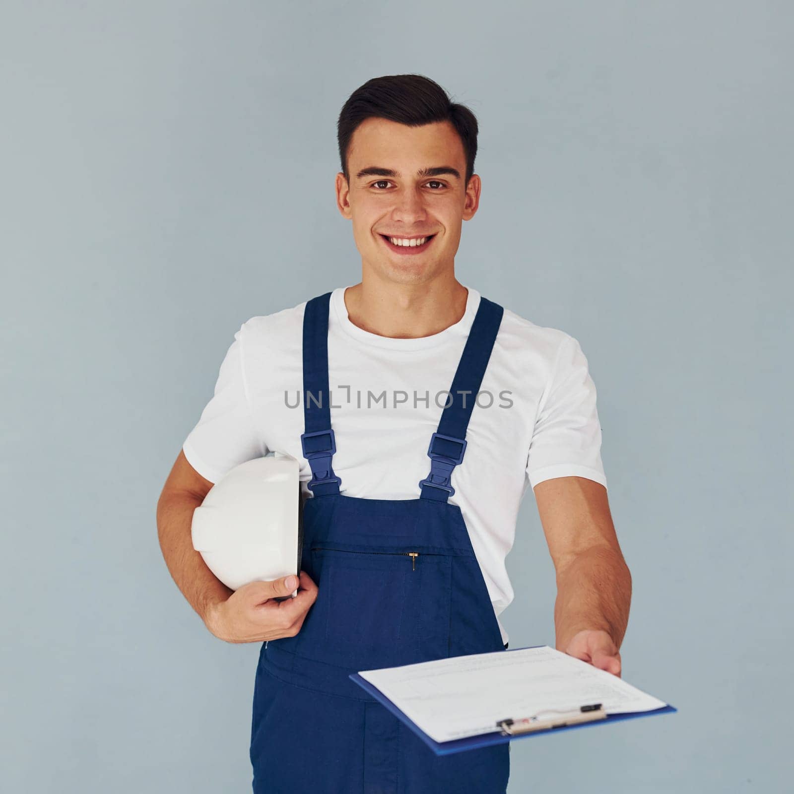 With notepad and hard hat. Male worker in blue uniform standing inside of studio against white background.