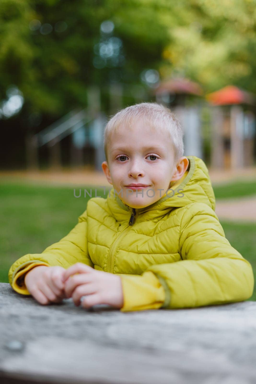 A boy sits at a table in the park in the fall season. Child Boy Son In Autumn Park, Sitting On Wooden Bench And Table. Little Kid Outdoors