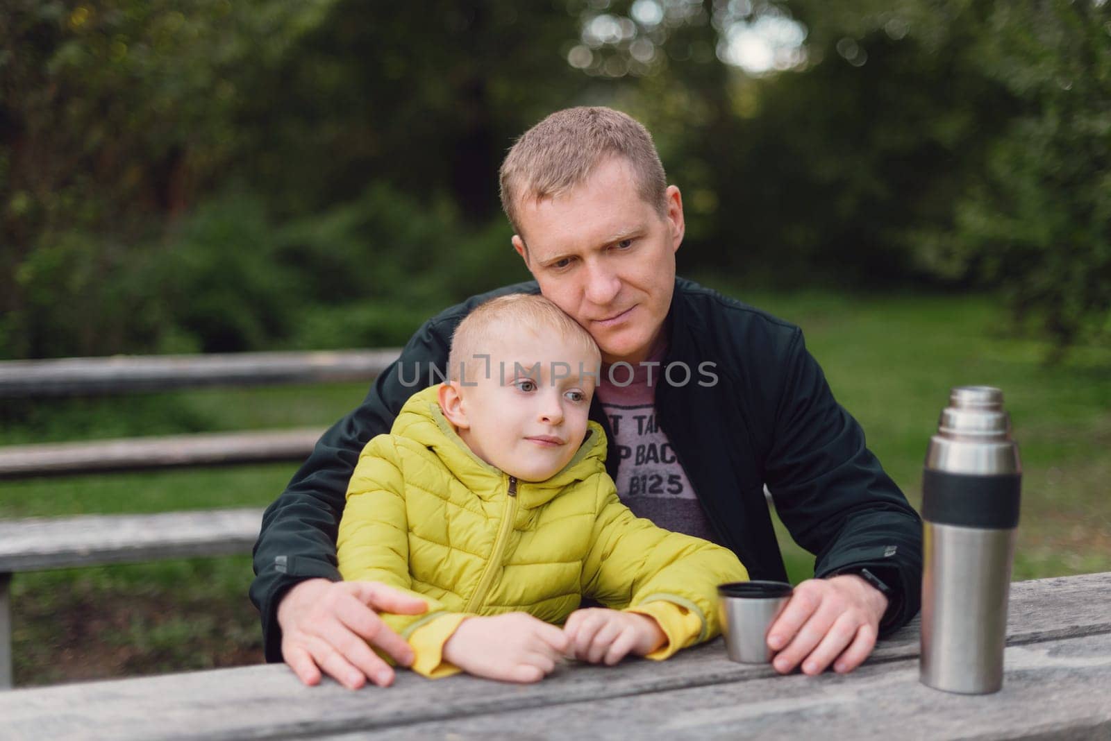 Happy family: father and child boy son playing and laughing in autumn park, sitting on wooden bench. Father and little kid having fun outdoors, playing together. Father and son sitting on a bench and talking. dad son park bench table autumn thermos.