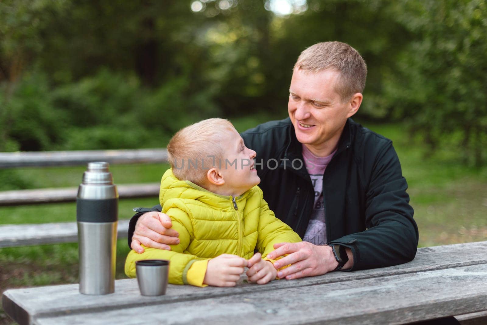 Happy Family: Father And Child Boy Son Playing And Laughing In Autumn Park, Sitting On Wooden Bench And Table. Father And Little Kid Having Fun Outdoors, Playing Together. Father And Son Sitting On A Bench And Talking. Thermos by Andrii_Ko