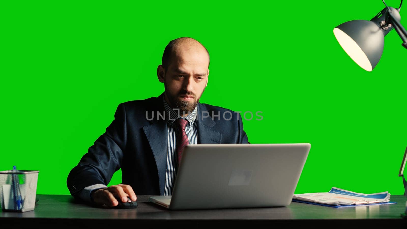 Businessman working on laptop over greenscreen backdrop, using documents at desk. Successful team leader in suit posing over isolated chroma key template, blank copyspace background.