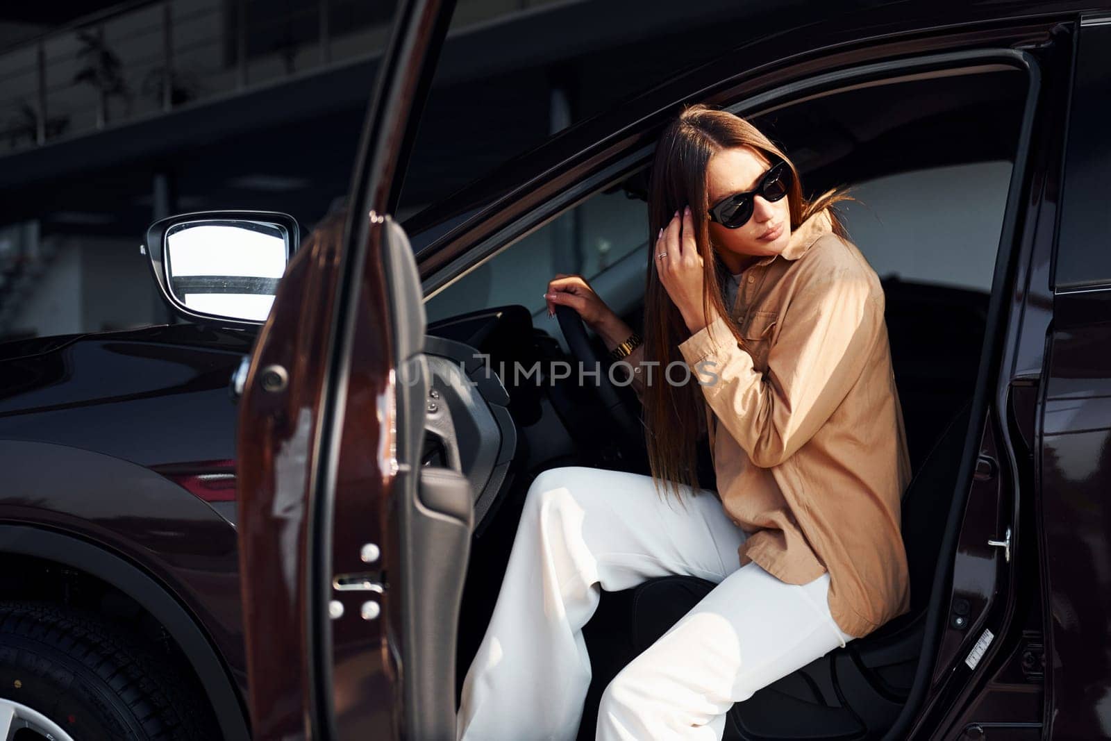 Sits with car's window opened. Fashionable beautiful young woman and her modern automobile.