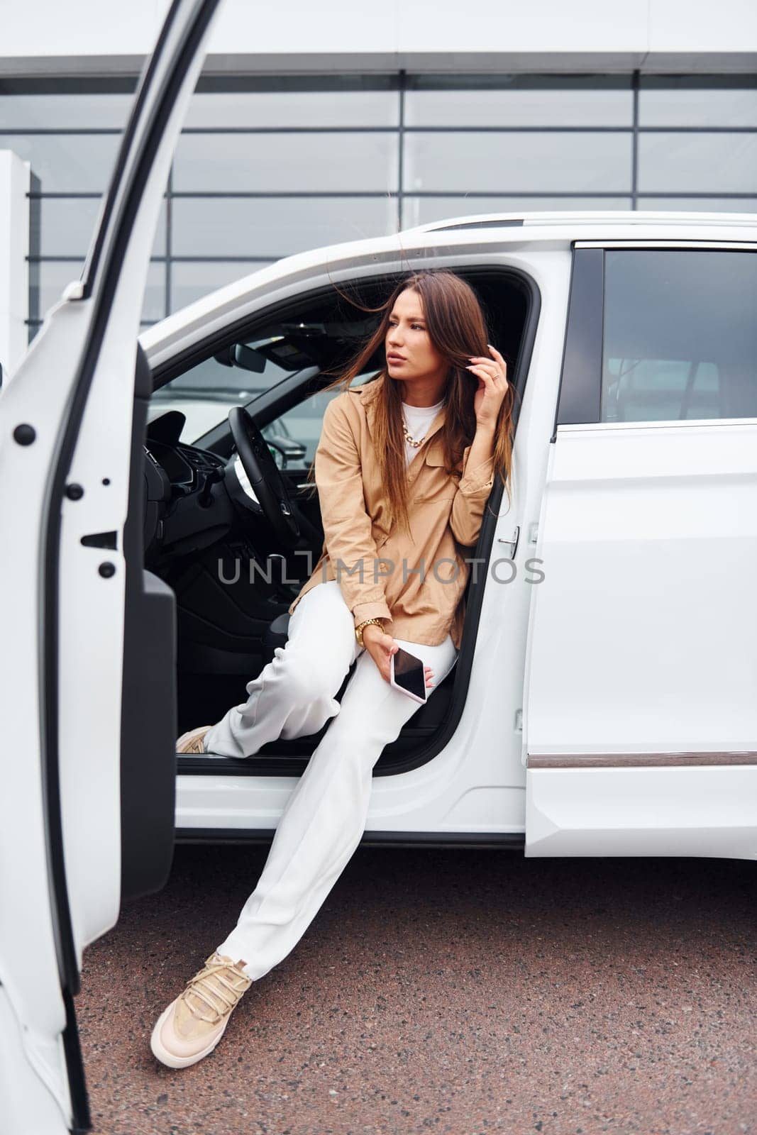 Outdoors against modern building. Fashionable beautiful young woman and her modern automobile.