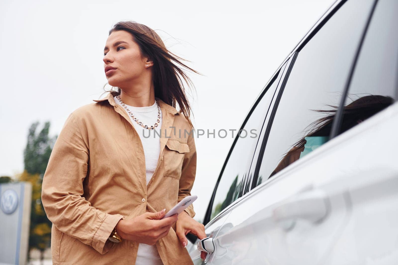 Standing outside and holds phone. Fashionable beautiful young woman and her modern automobile.