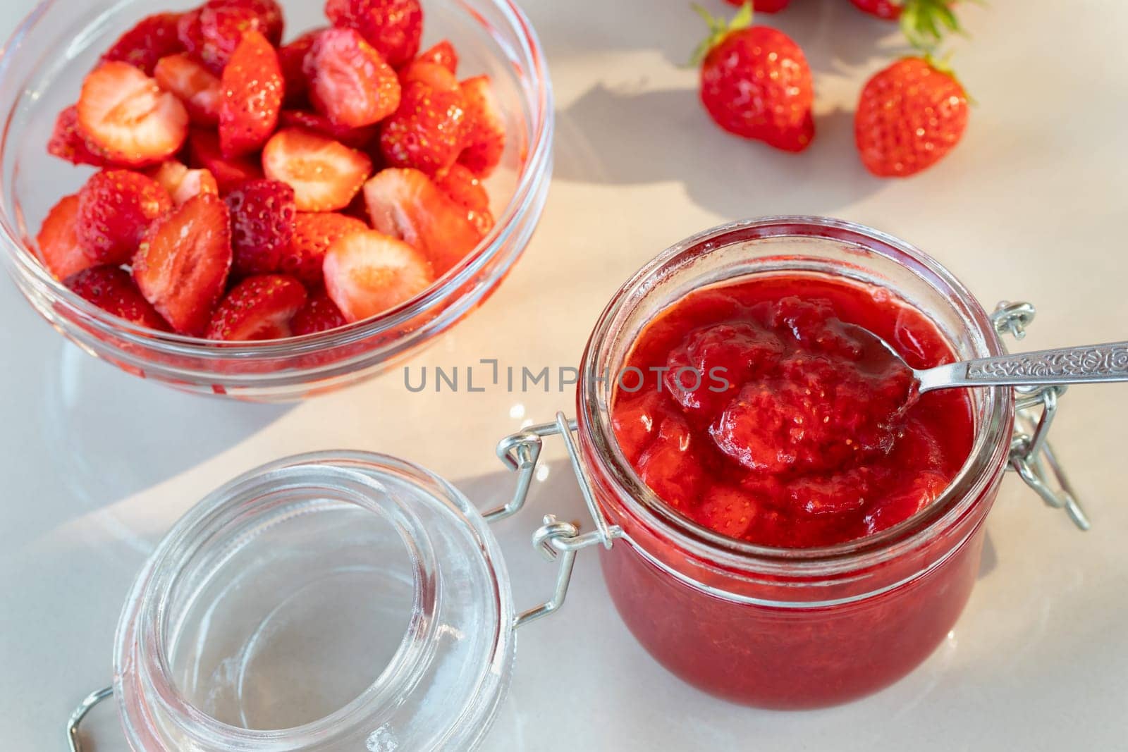 Glass jar with strawberry jam prepared for canning and fresh strawberries in a bowl on the table.