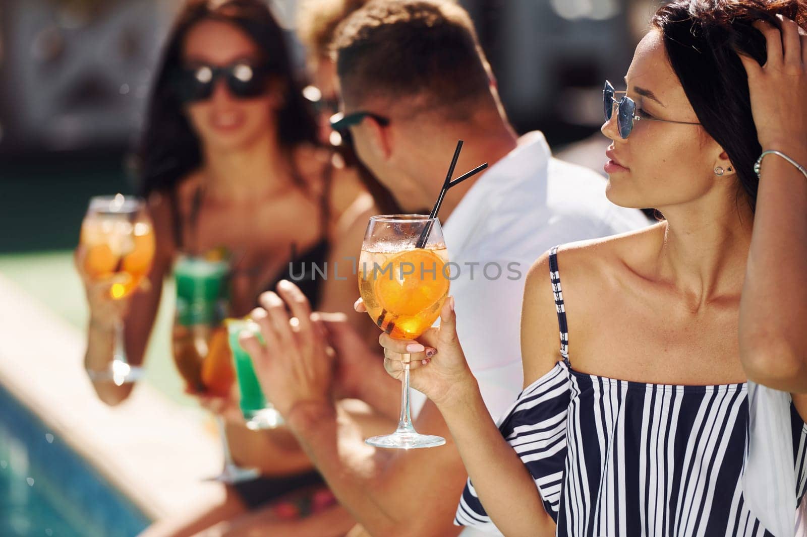 Fresh cocktails in hands. Group of young happy people have fun in swimming pool at daytime.