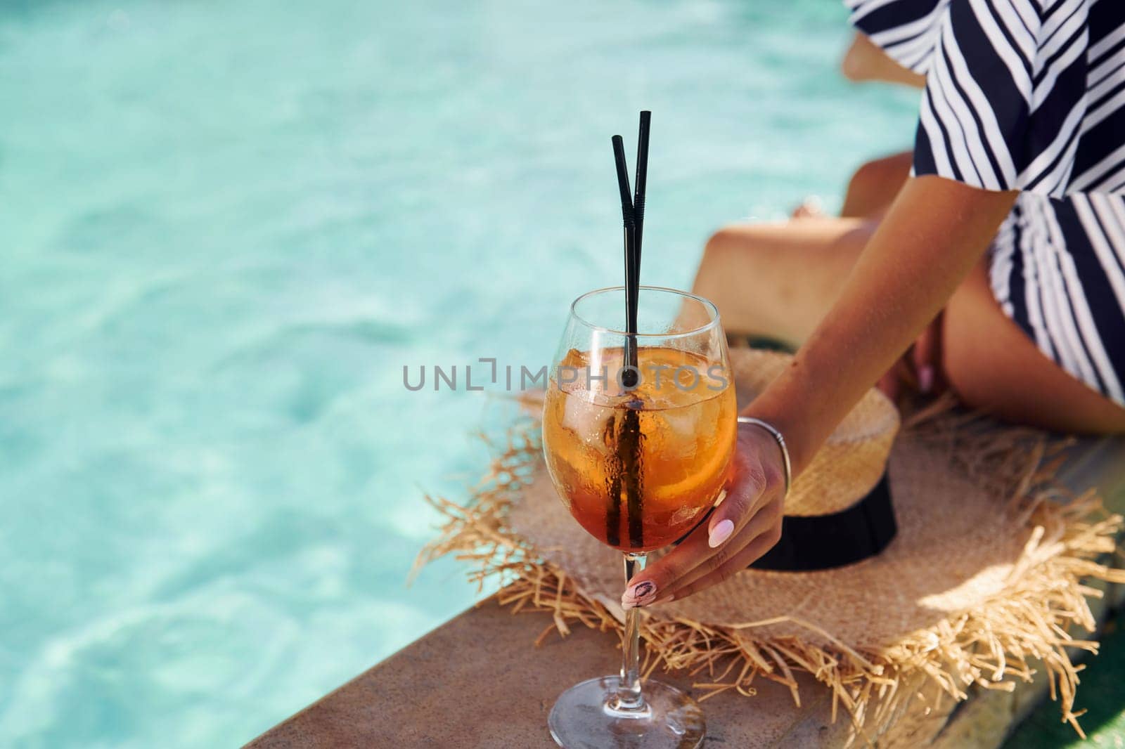 Young woman sits near swimming pool at daytime with cocktail by Standret