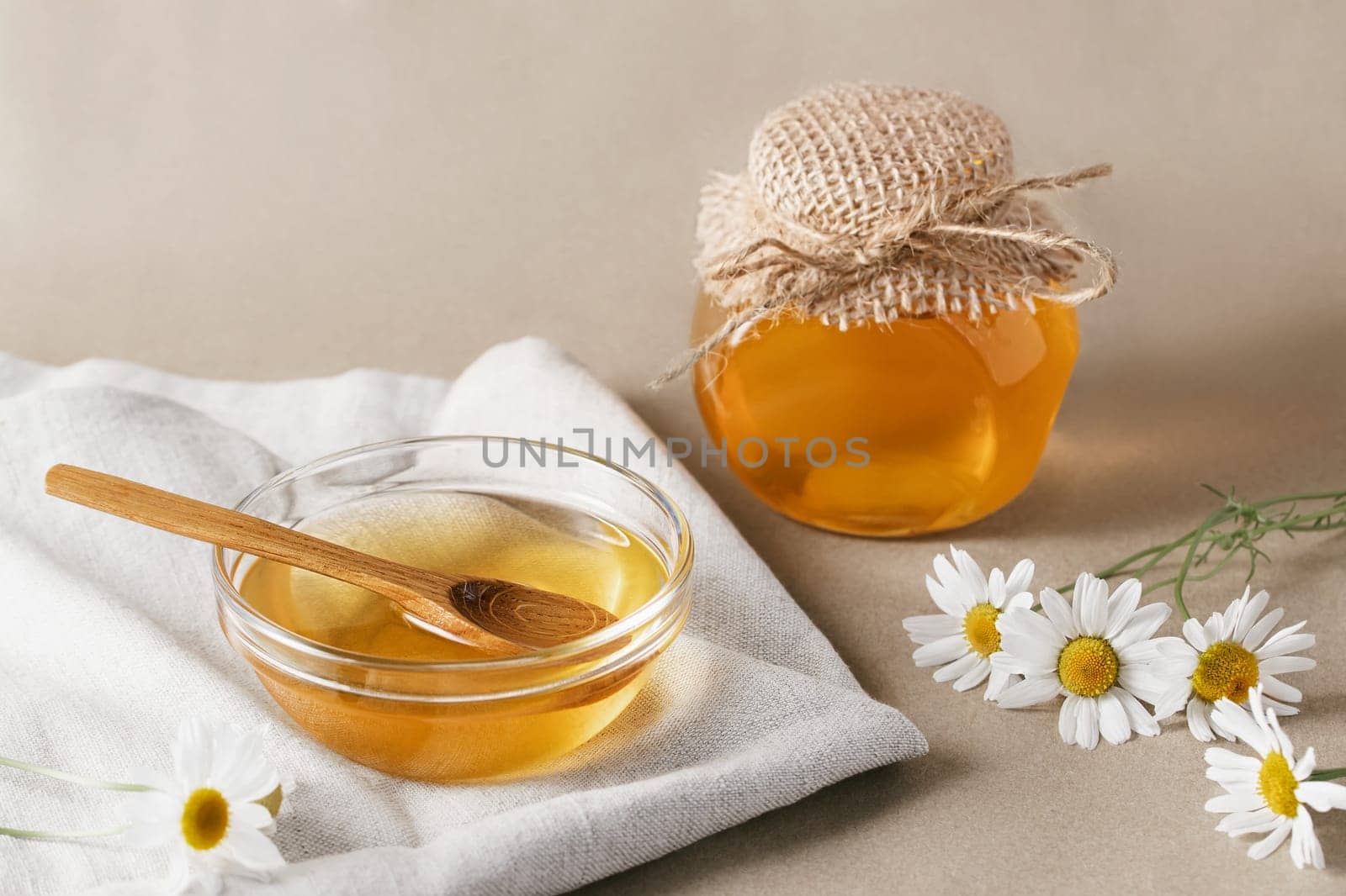 Chamomile syrup in a small bowl and in a jar and chamomile flowers on a linen kitchen towel.