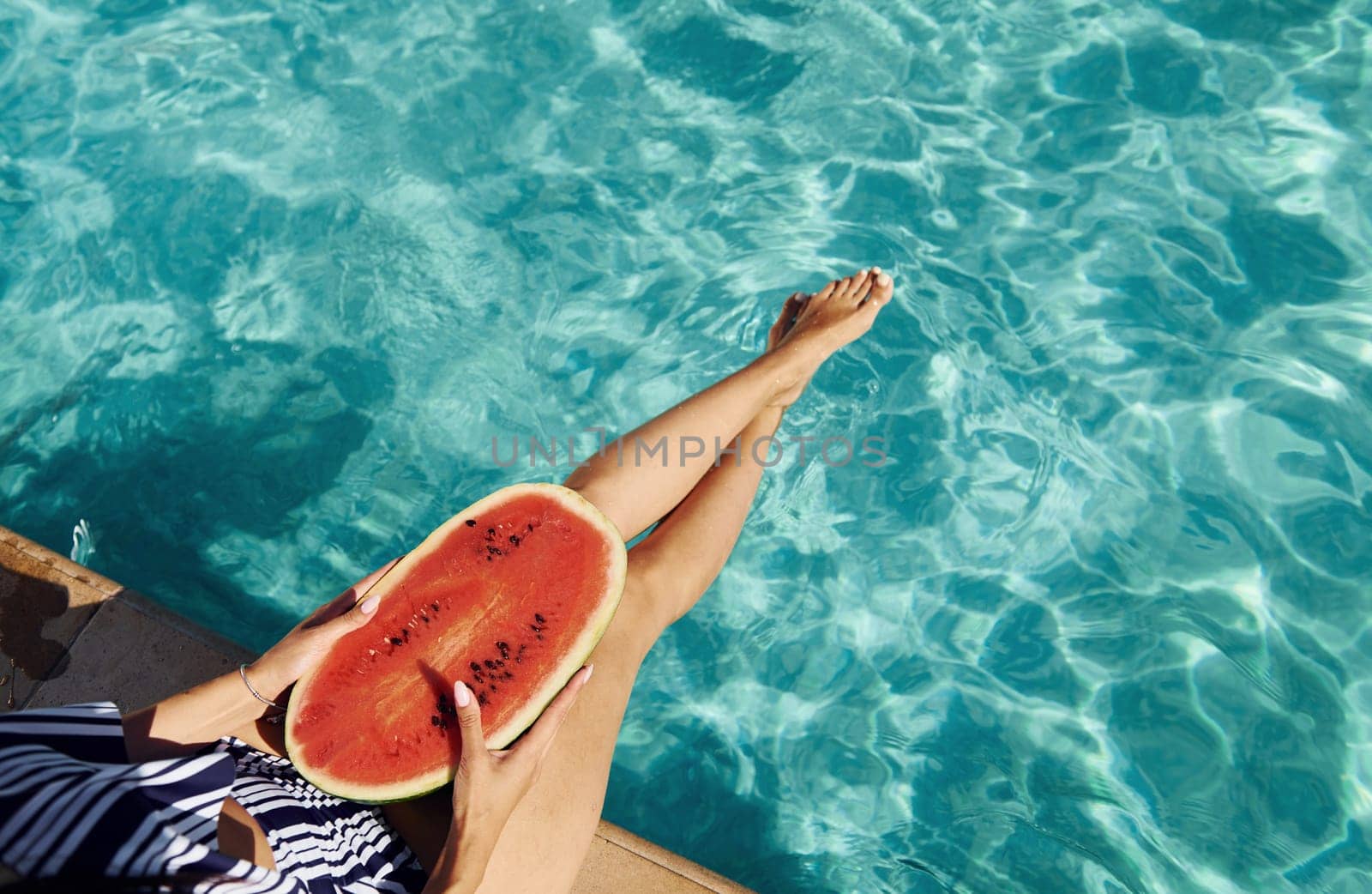 Young woman sits near swimming pool at daytime with watermelon.