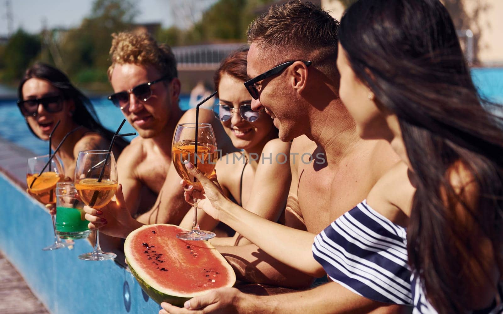 Holding watermelon. Group of young happy people have fun in swimming pool at daytime.