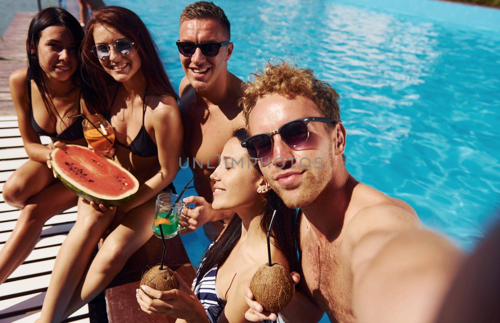 Holding watermelon. Group of young happy people have fun in swimming pool at daytime by Standret