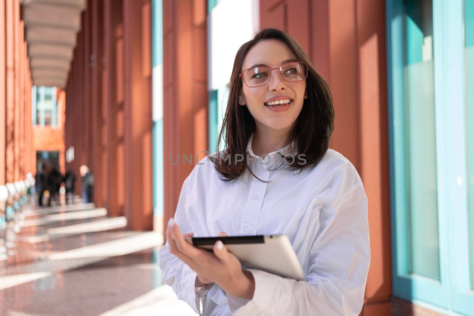 Businesswoman in glasses using digital tablet touching screen looking away, standing outdoor dressed white shirt. Female freelancer holding tablet while walk outside. Copy space