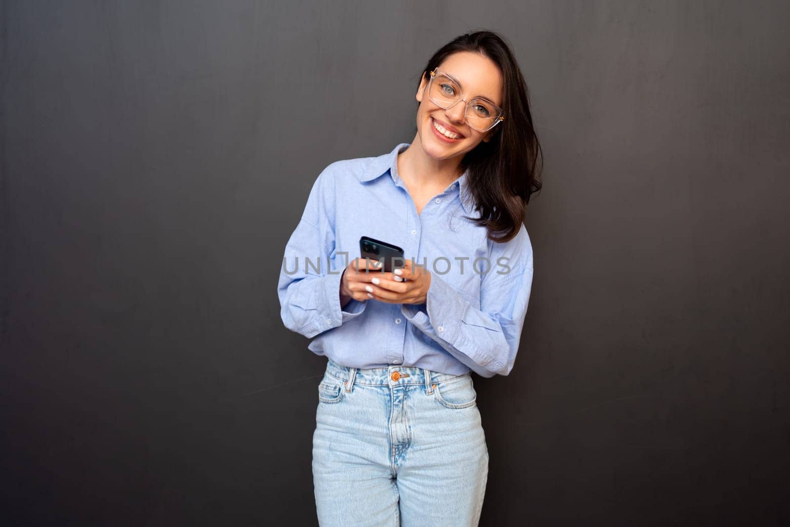 Positive smiling woman holding smartphone in hand looking at camera, wear glasses, types text message on mobile phone, enjoys online communication, on black studio background. Technology concept