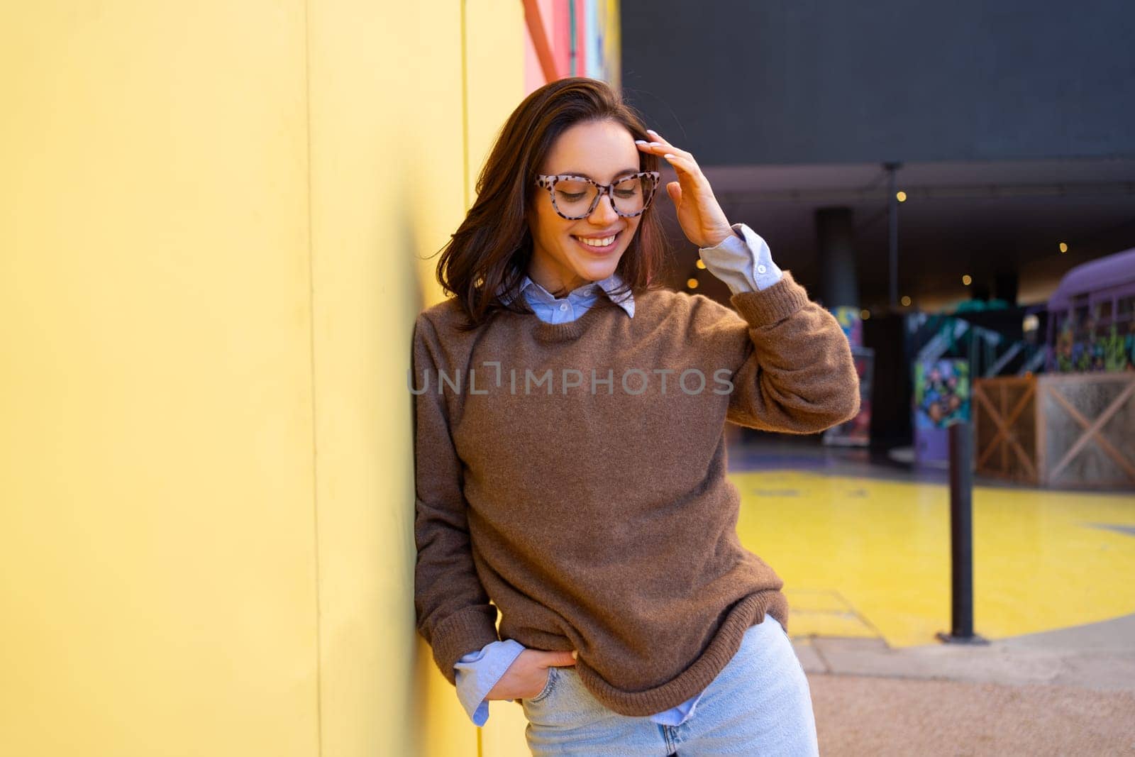 Happy woman in glasses outdoor leaning on yellow wall. Positive people concept. Smiling girl looking down, hands in pocket, dressed sweater and jeans, correct hairstyle