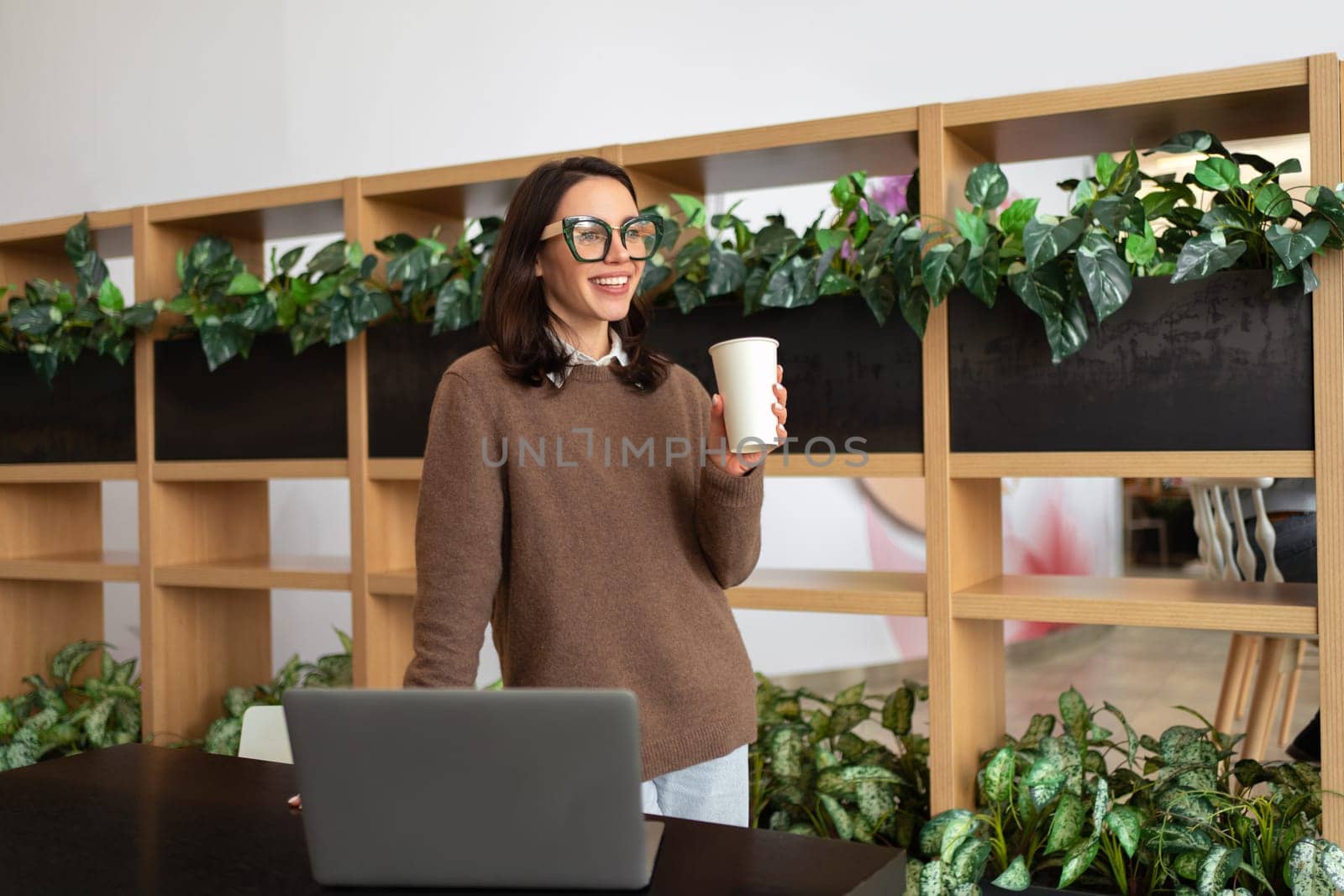 Happy European Female Entrepreneur Standing Near Desk With Laptop Computer Posing Smiling In Modern Office Indoor. Freelance Career And Successful Entrepreneurship Concept