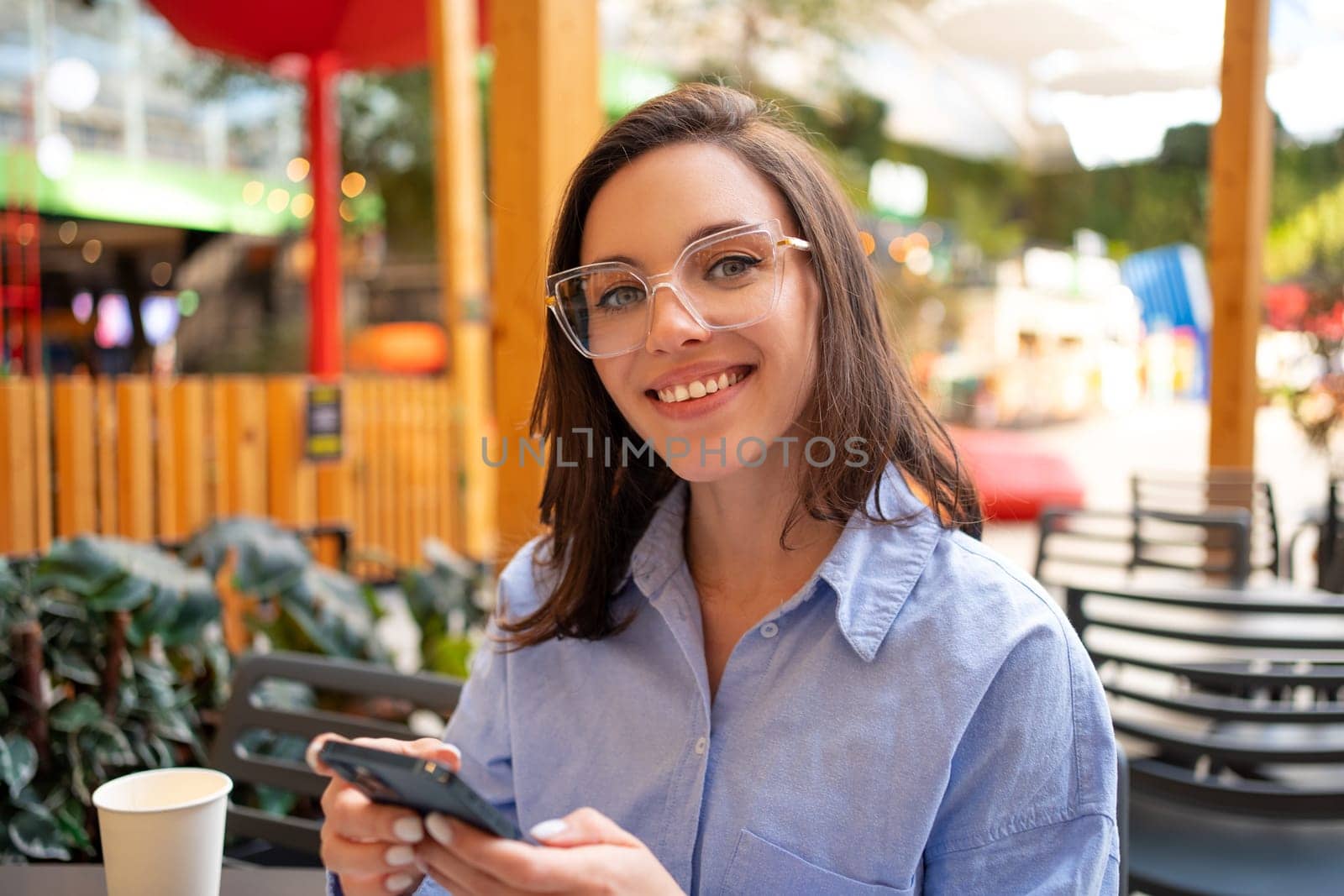 Portrait woman using smartphone while enjoying summer on outdoor terrace cafe or coffee shop, looking at camera and smile. Female person wear glasses showing teeth smile holding mobile phone in hand