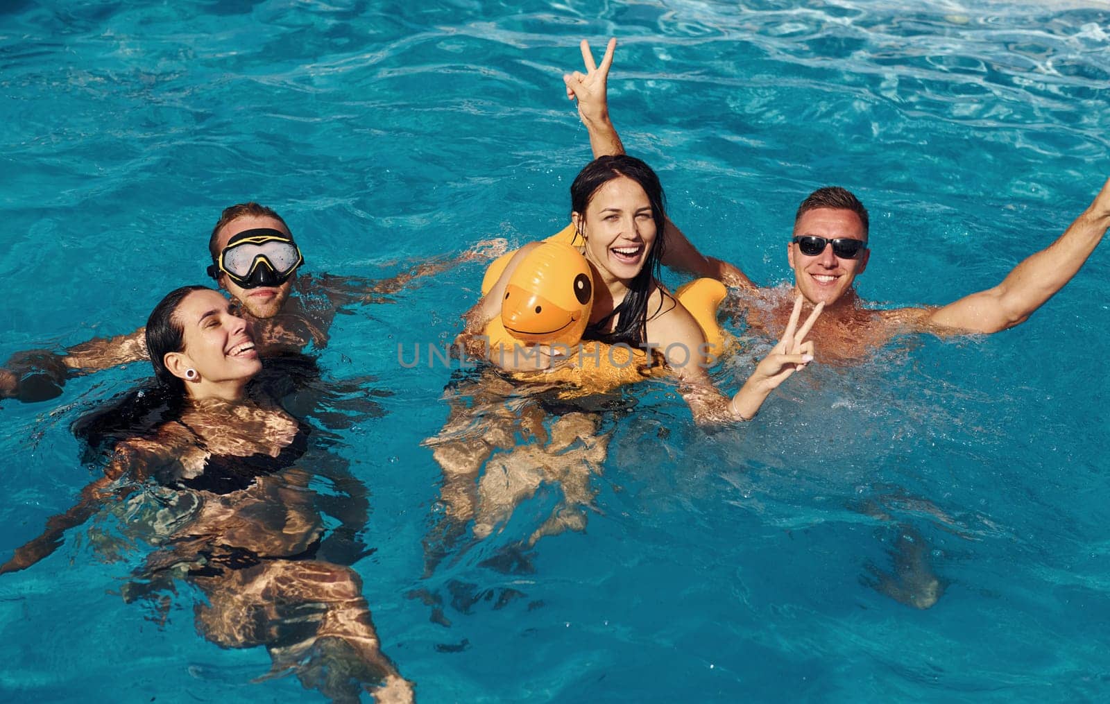 Using yellow colored duck to swim. Group of young happy people have fun in swimming pool at daytime.