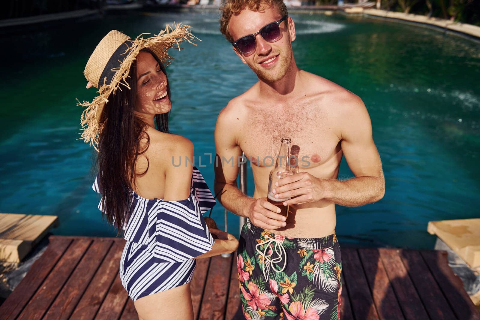 Holding drinks in hands. Cheerful couple or friends together in swimming pool at vacation by Standret