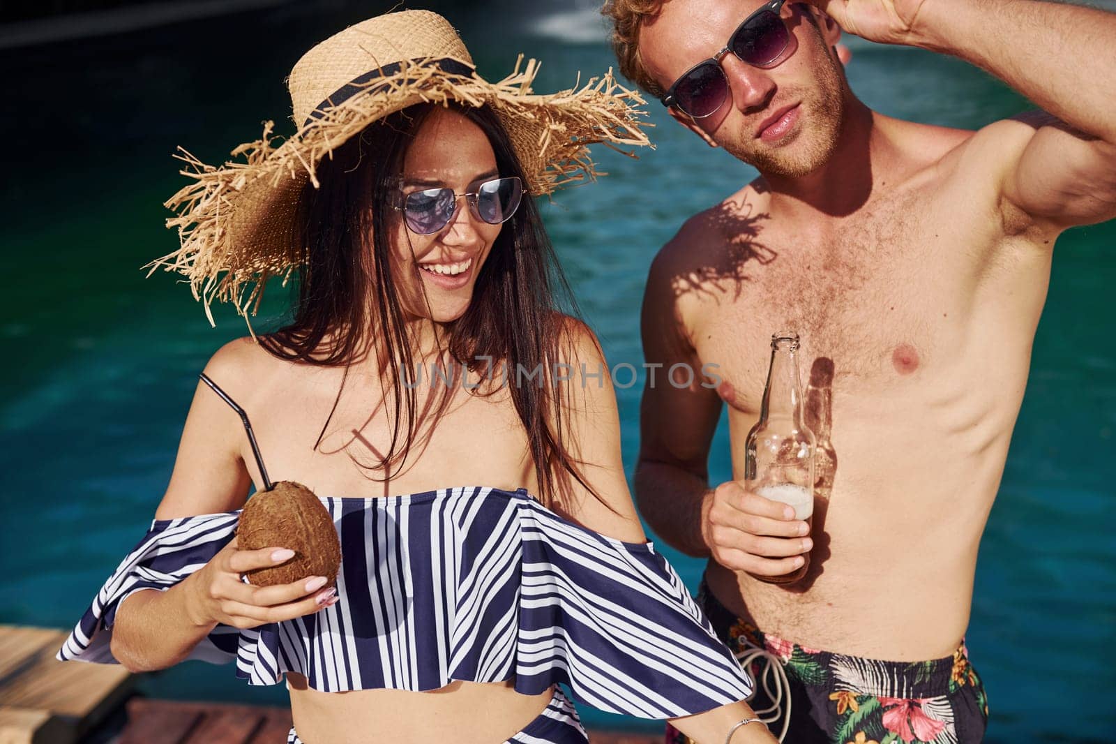 Holding drinks in hands. Cheerful couple or friends together in swimming pool at vacation by Standret