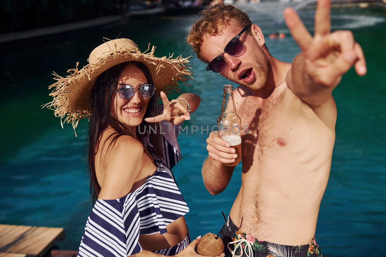 Holding drinks in hands. Cheerful couple or friends together in swimming pool at vacation by Standret