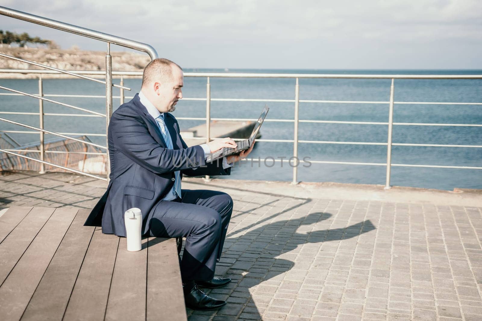 Confident middle age businessman working remotely online, typing on a laptop keyboard while sitting on a beach at sunset. Working remotely on vacation, running an online business from a distance. by panophotograph