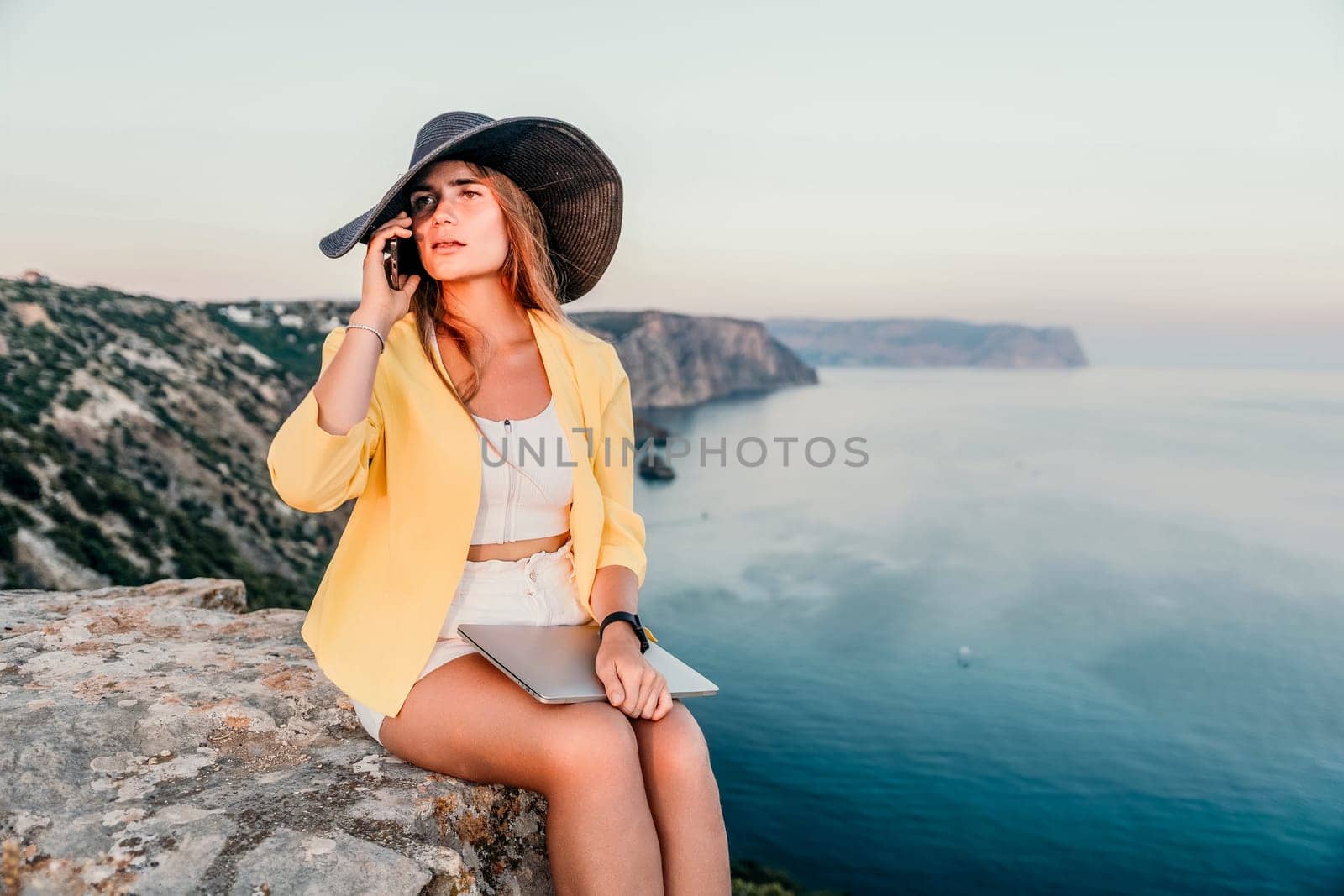 Successful business woman in yellow hat working on laptop by the sea. Pretty lady typing on computer at summer day outdoors. Freelance, travel and holidays concept.