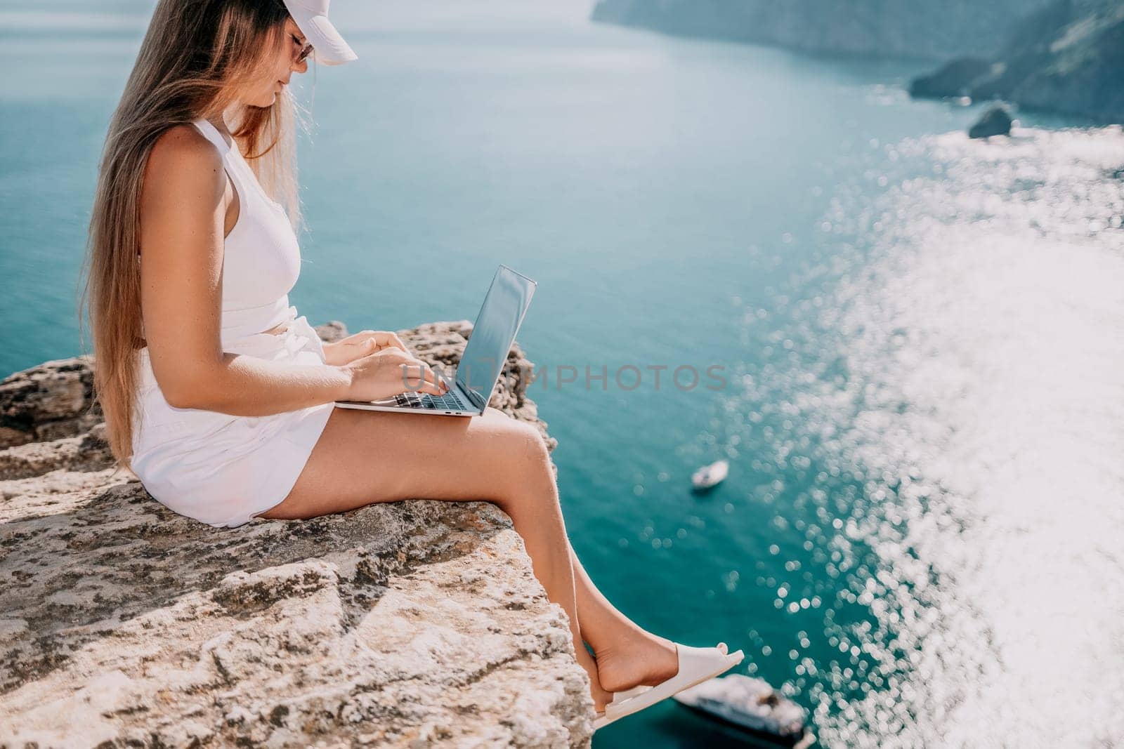 Successful business woman in yellow hat working on laptop by the sea. Pretty lady typing on computer at summer day outdoors. Freelance, travel and holidays concept.