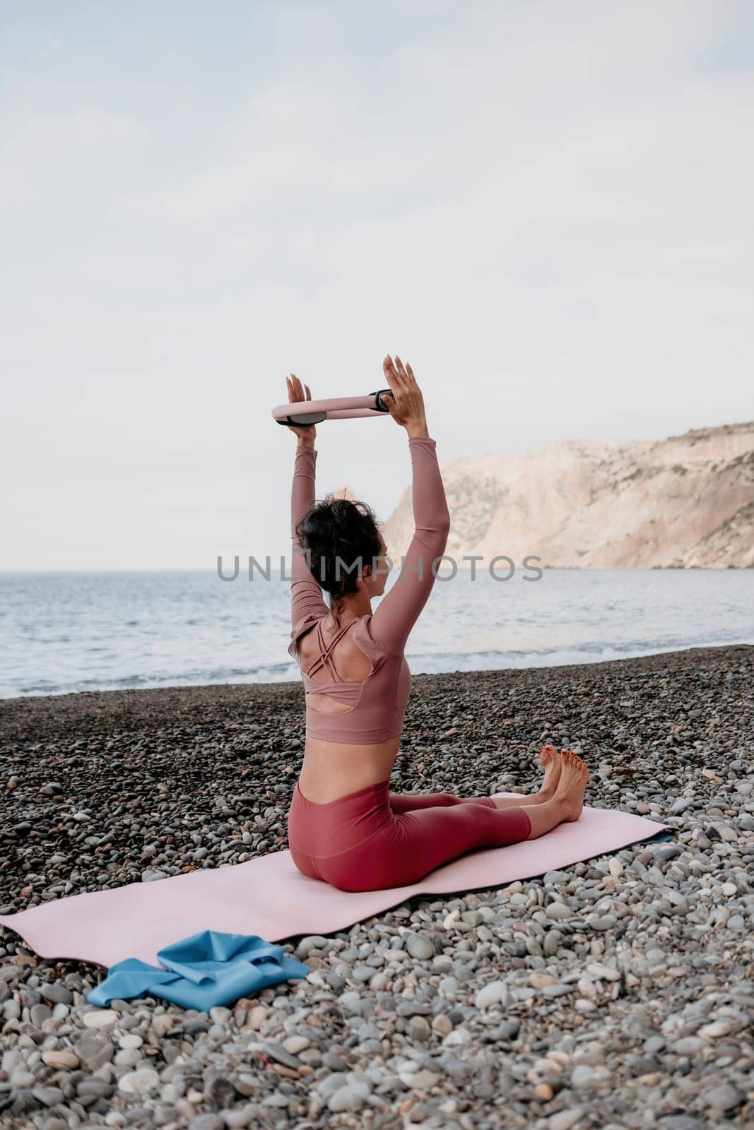 Middle aged well looking woman with black hair doing Pilates with the ring on the yoga mat near the sea on the pebble beach. Female fitness yoga concept. Healthy lifestyle, harmony and meditation.