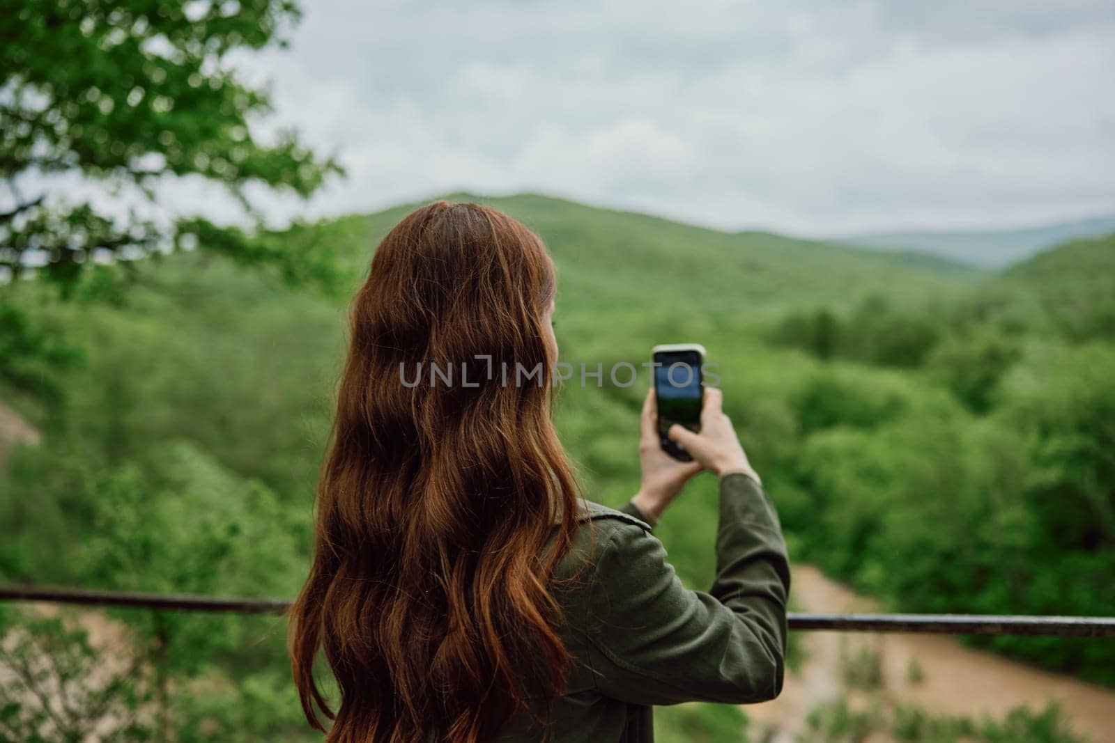 a woman in a raincoat takes pictures on the phone of a beautiful view of the forest. Travel, technology, mobile photography. High quality photo