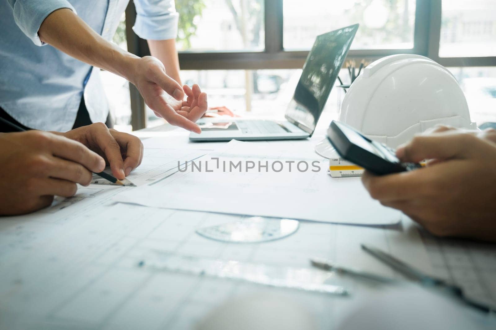Close-up hands shot of three engineer, developers, or architects working together, drawing graphic design of construction project, and discussing for improvement at desk with papers, compass, helmet, calculator, rulers, and laptop. Engineering and Teamwork concept.