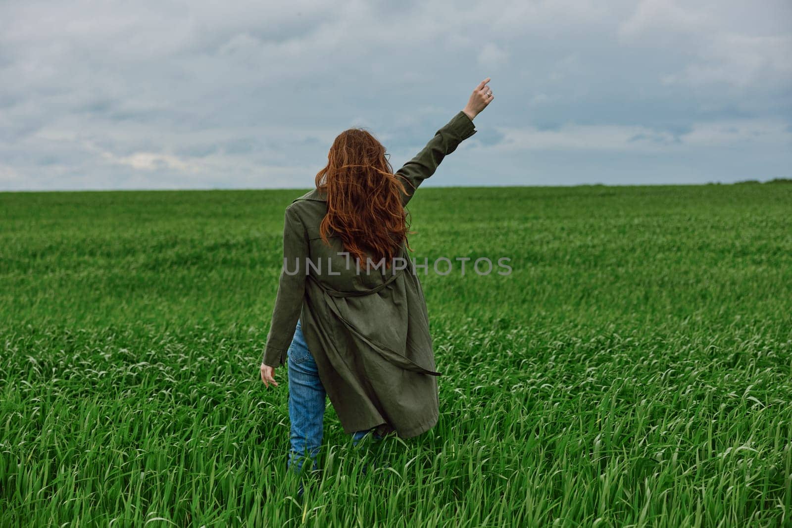 a red-haired woman stands with her back to the camera in a long coat with her hand raised up in a green field in rainy weather by Vichizh