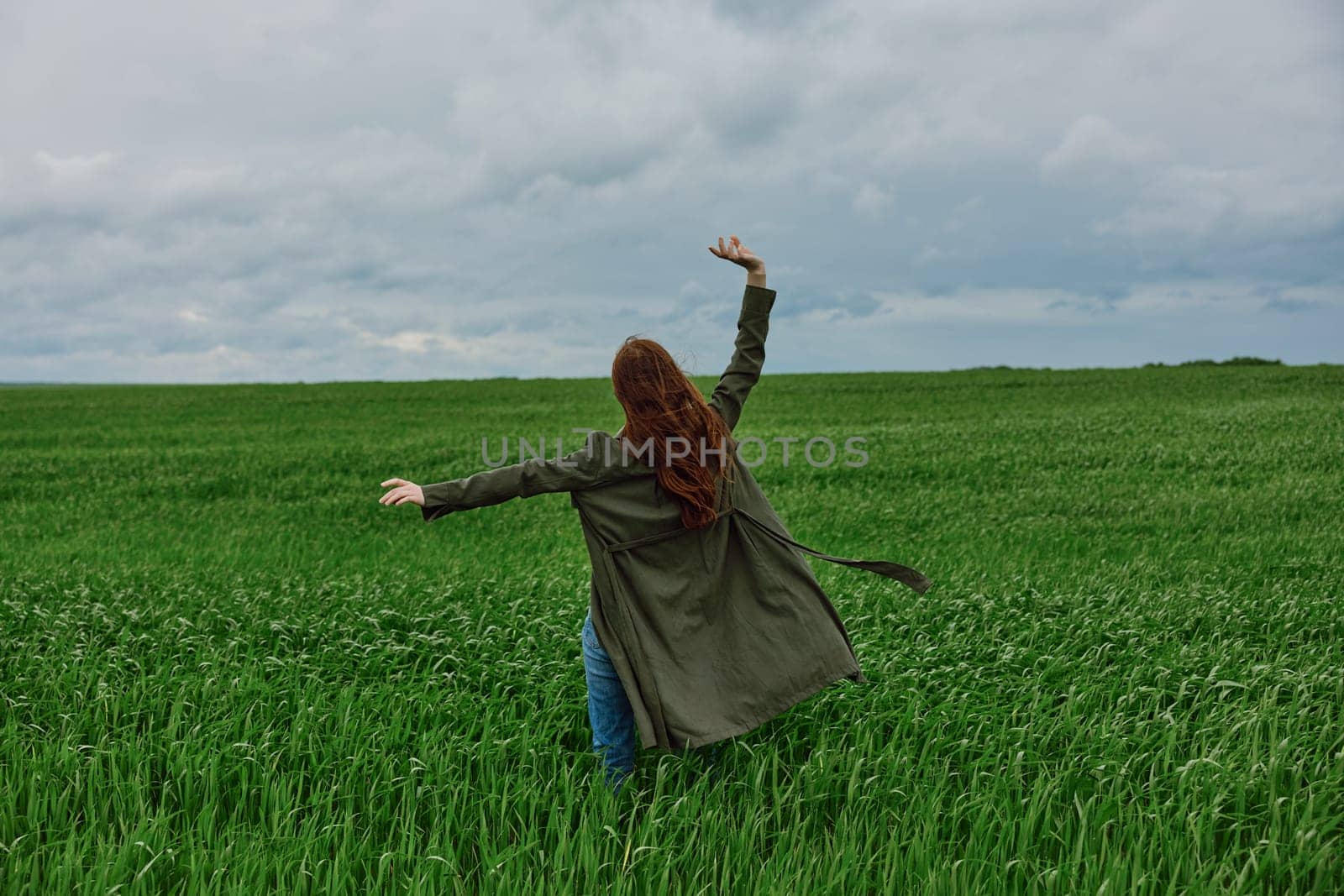 a red-haired woman stands with her back to the camera in a long coat with her hand raised up in a green field in rainy weather. High quality photo