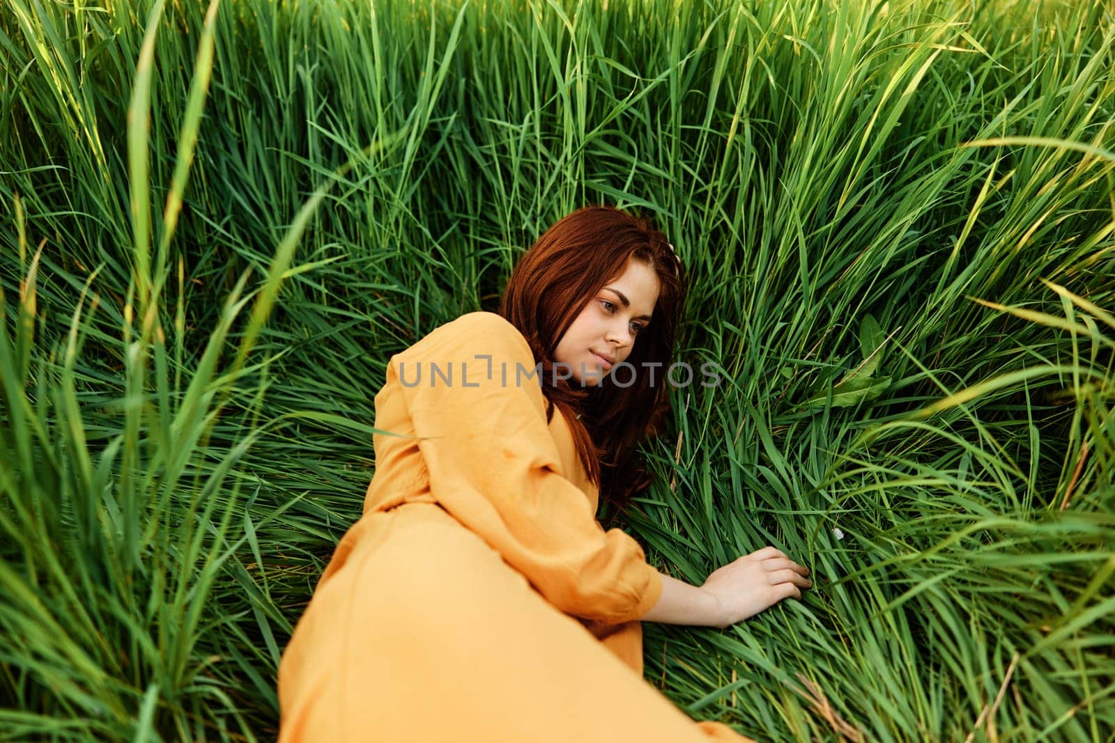 a happy woman is lying in the tall green grass in a long orange dress and smiling pleasantly looks away holding the leaves with her hand, enjoying nature. Close horizontal photo by Vichizh
