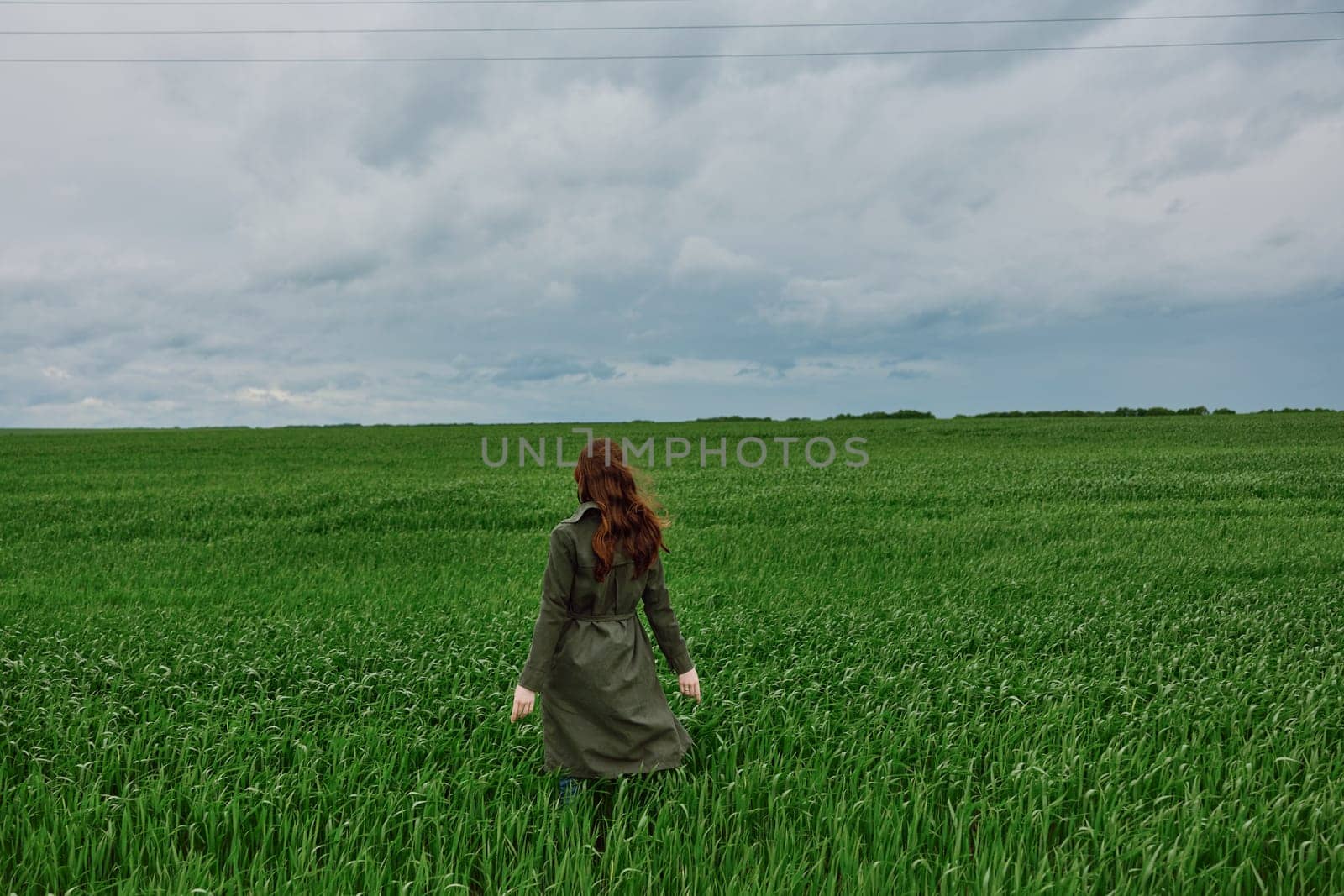 a red-haired woman in a long raincoat stands in a green field in cloudy weather with her back to the camera enjoying nature by Vichizh