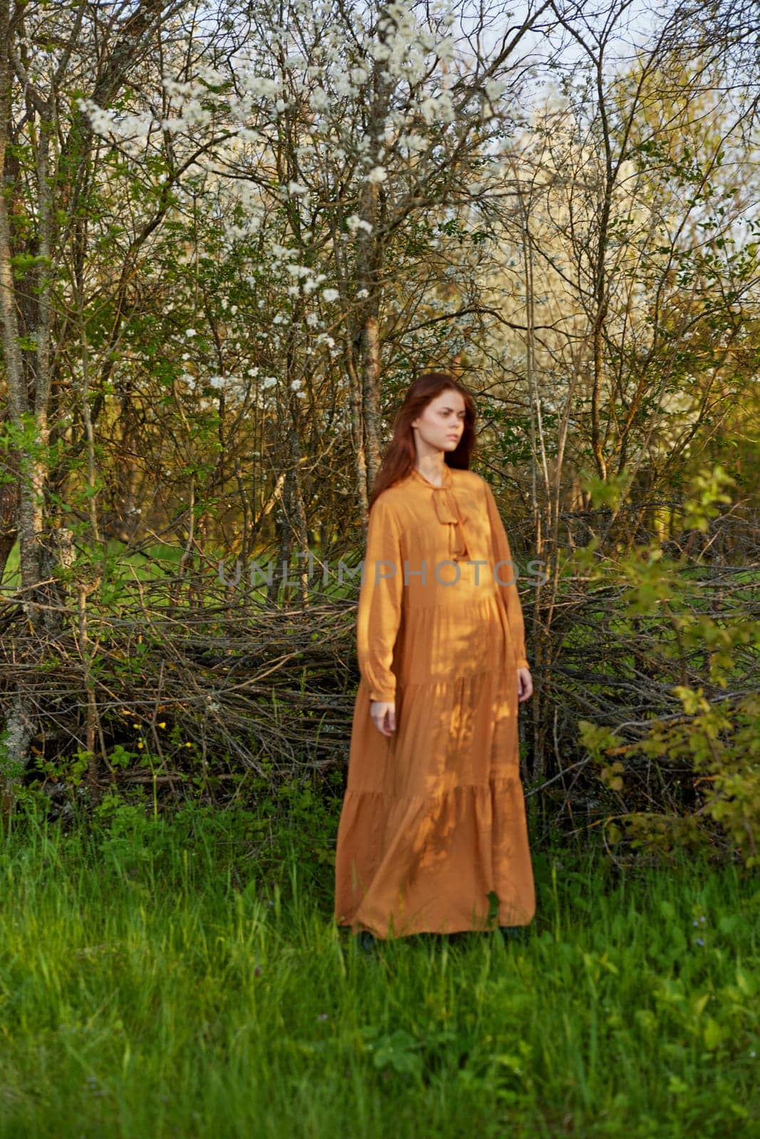 an elegant, sophisticated woman poses relaxed standing near a wicker fence at the dacha in a long orange dress looking to the side. Vertical photography in nature by Vichizh