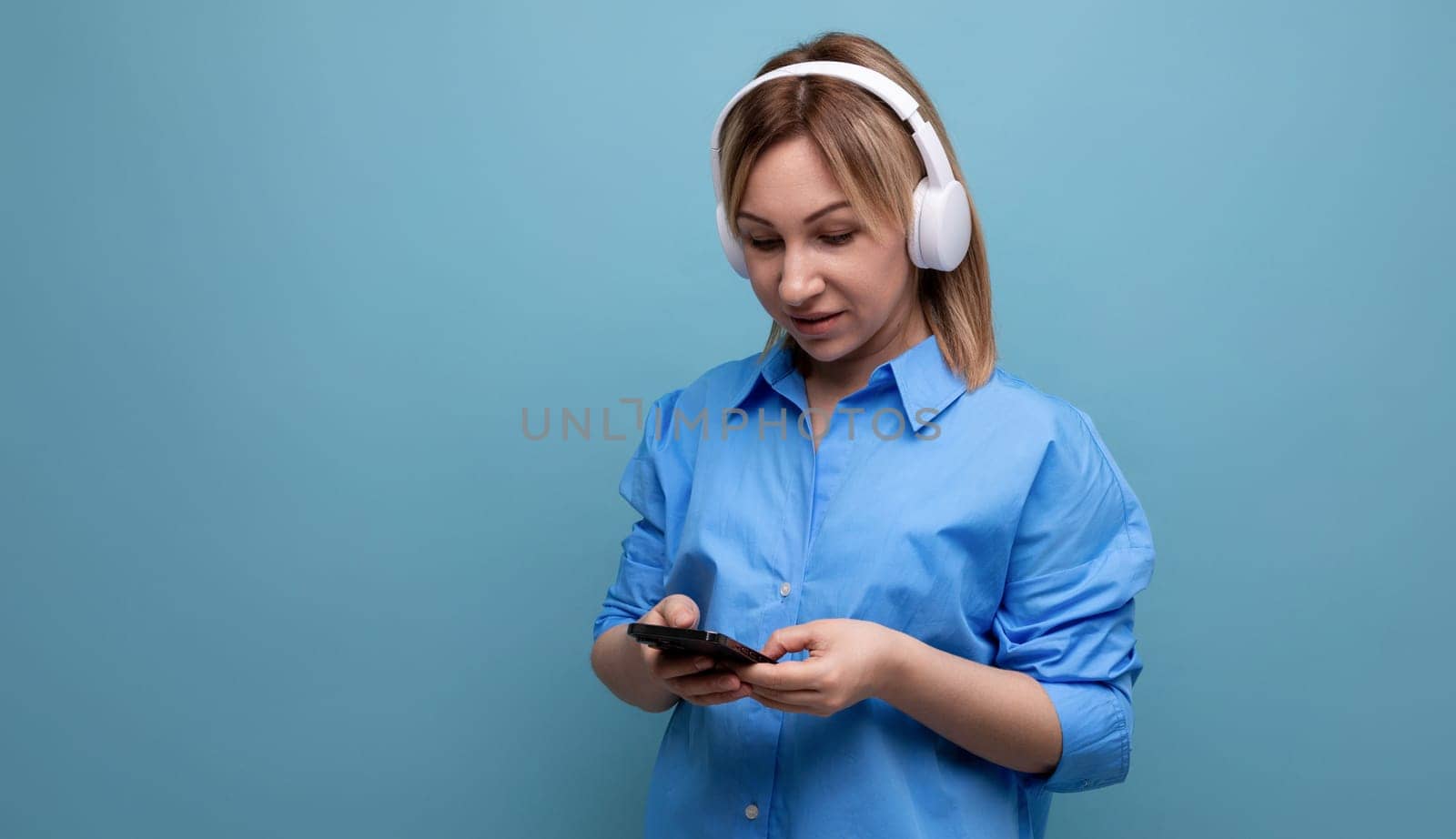 close-up of a millennial woman in a casual shirt with big white headphones with a smartphone in her hands on a blue isolated background with copy space by TRMK