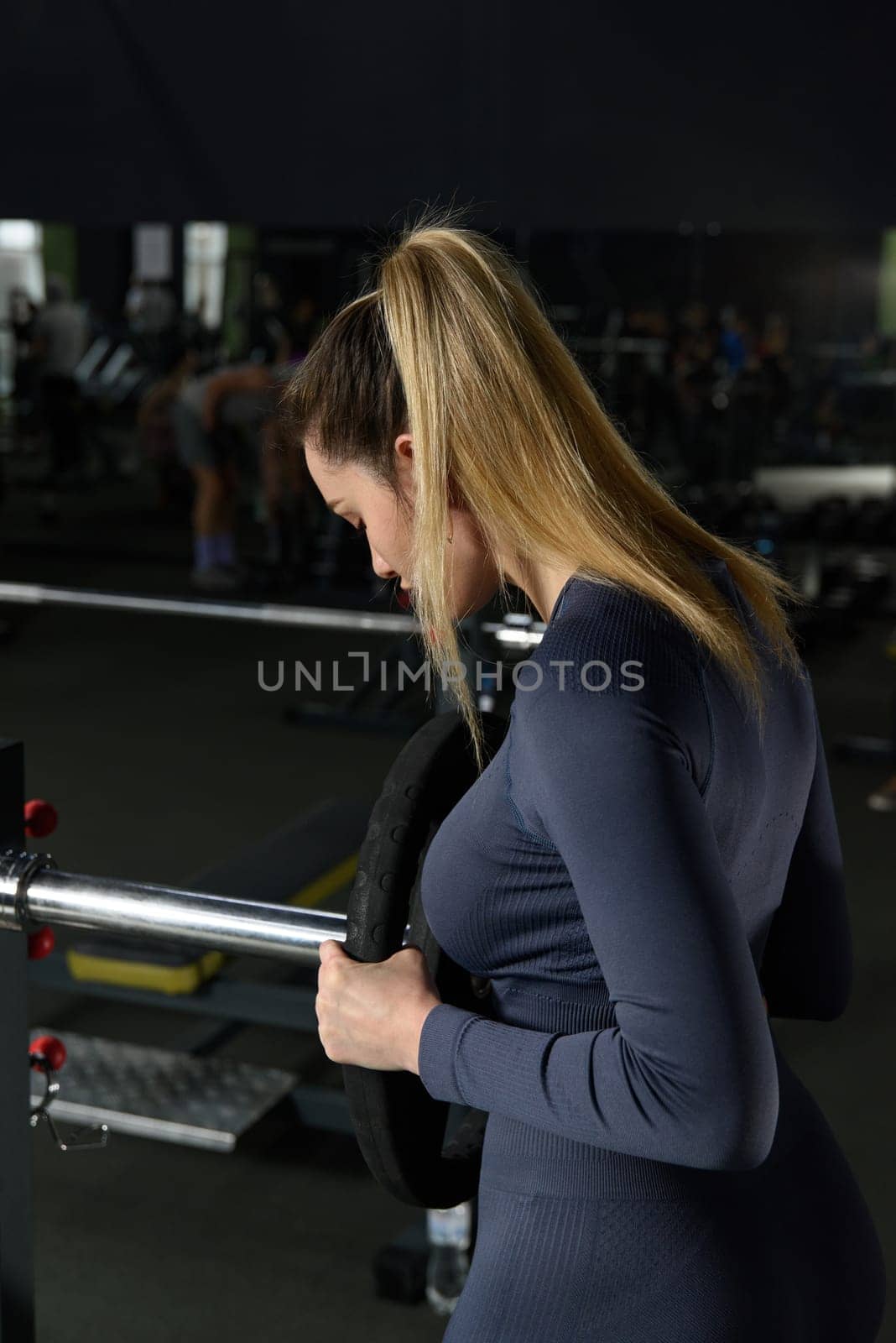 One adult caucasian woman female athlete putting weight plate on the barbel at gym. wearing blue leggins and long sleeve top