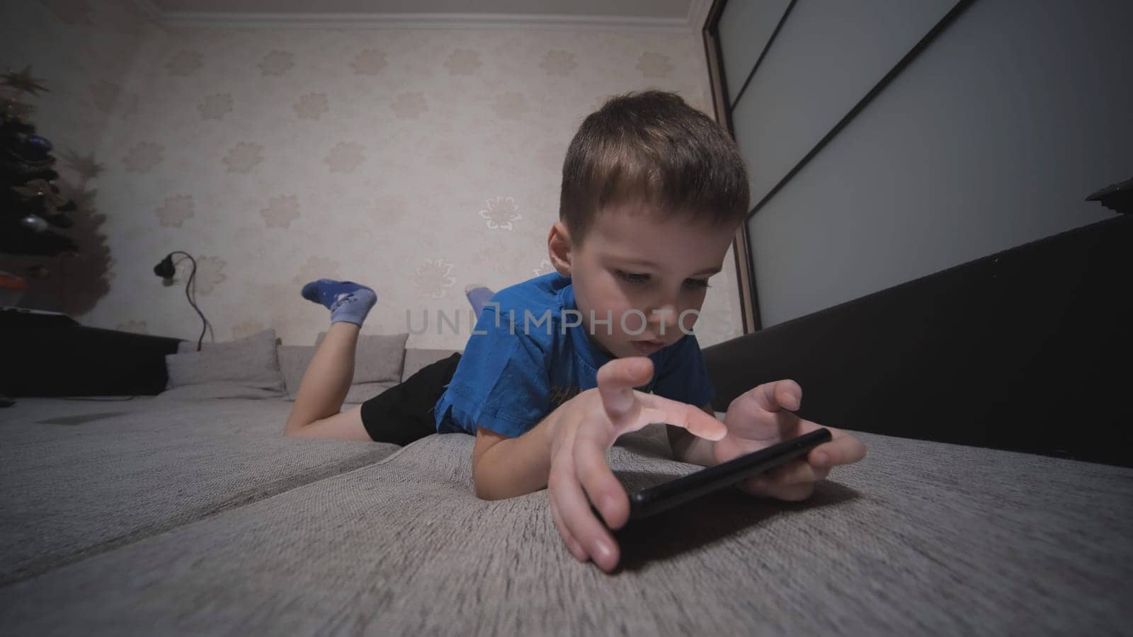 The boy plays on his smartphone while lying on the couch at home