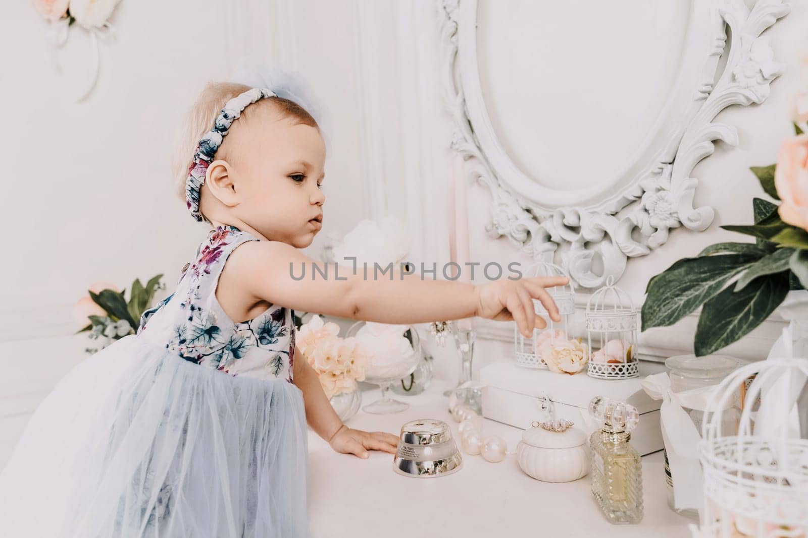 Baby girl elegant dress. A one-year-old girl in a puffy dress and a cute bow poses against the backdrop of a bright room with a dressing table and flowers. by Matiunina