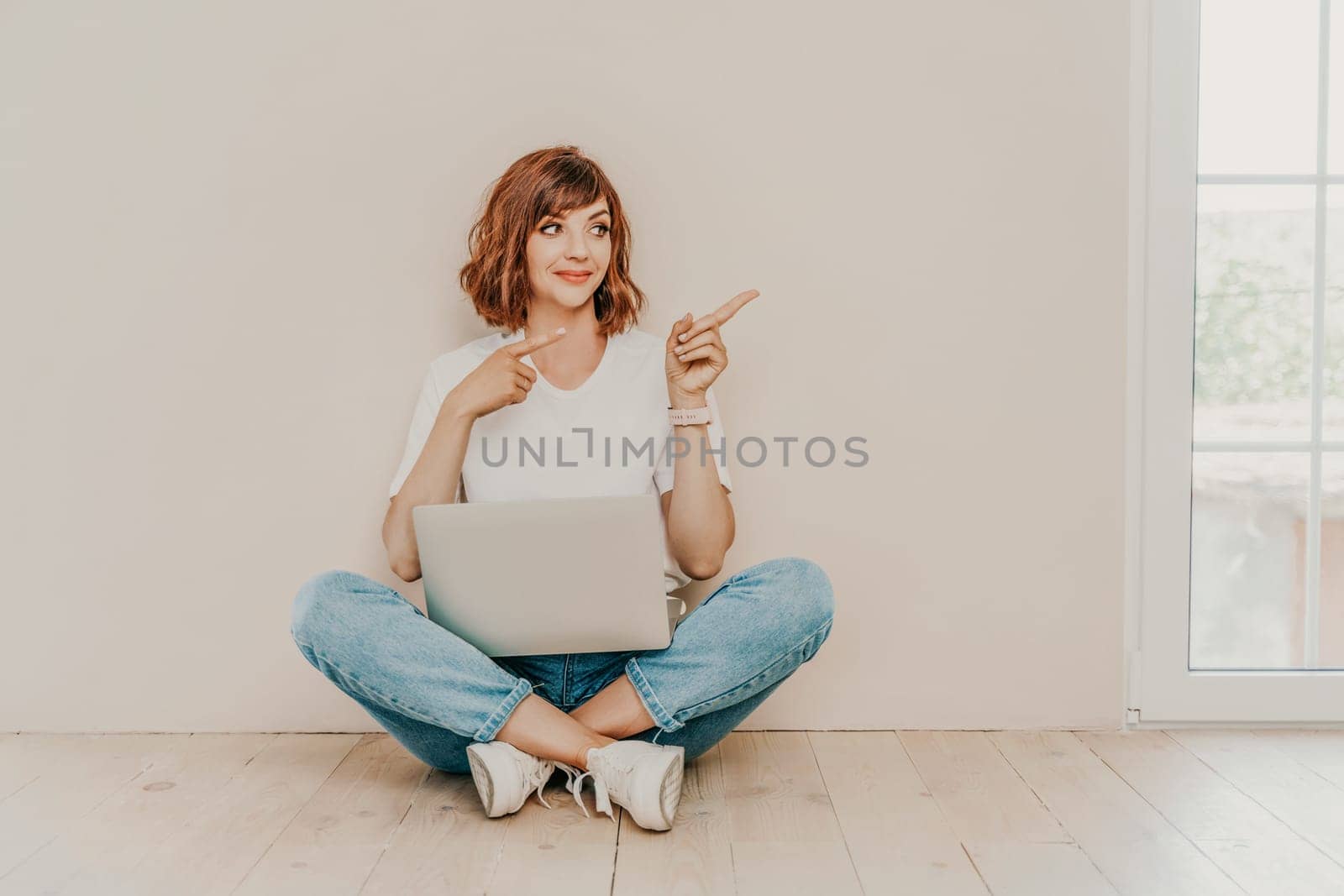 A brunette sits on the floor with a laptop on a beige wall background. She is wearing a white T-shirt, jeans and white sneakers