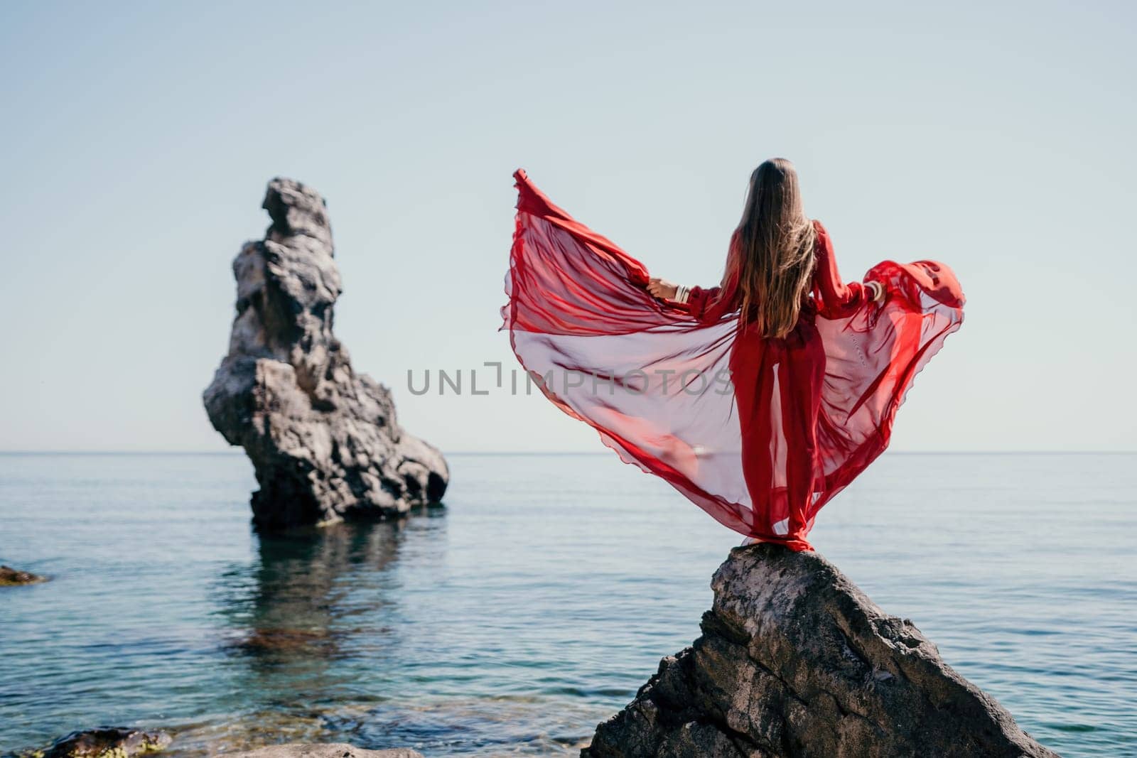 Woman travel sea. Young Happy woman in a long red dress posing on a beach near the sea on background of volcanic rocks, like in Iceland, sharing travel adventure journey by panophotograph