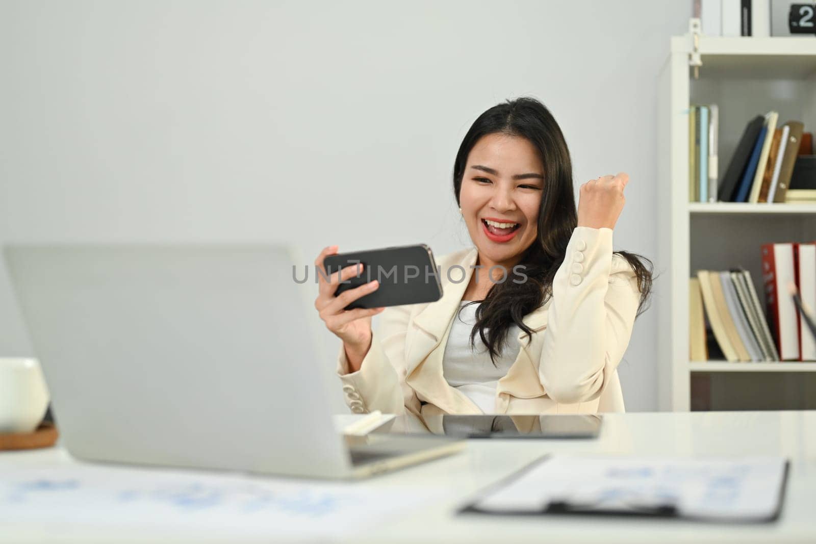 Overjoyed asian woman worker looking at mobile phone and celebrating success, excited by good news in email or message by prathanchorruangsak